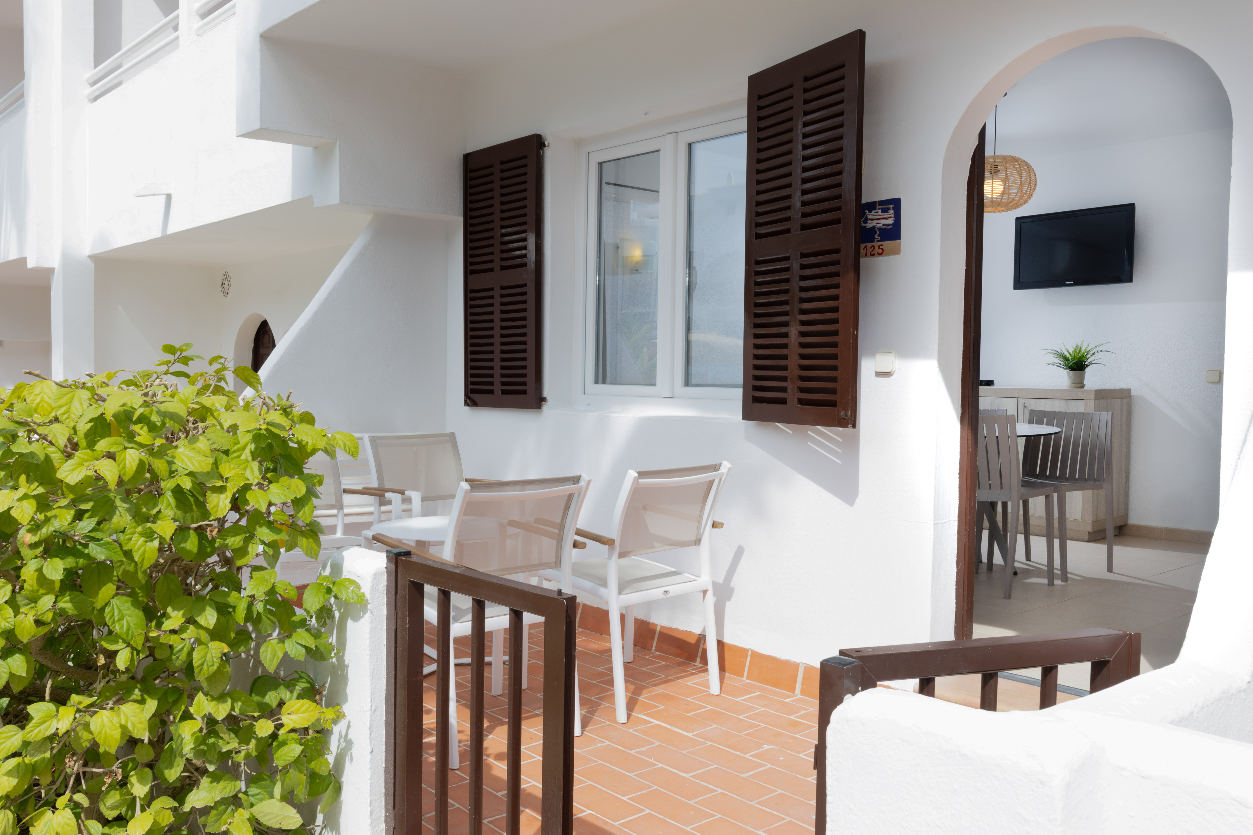 Bright apartment patio with outdoor seating, green bush, and room interior view.