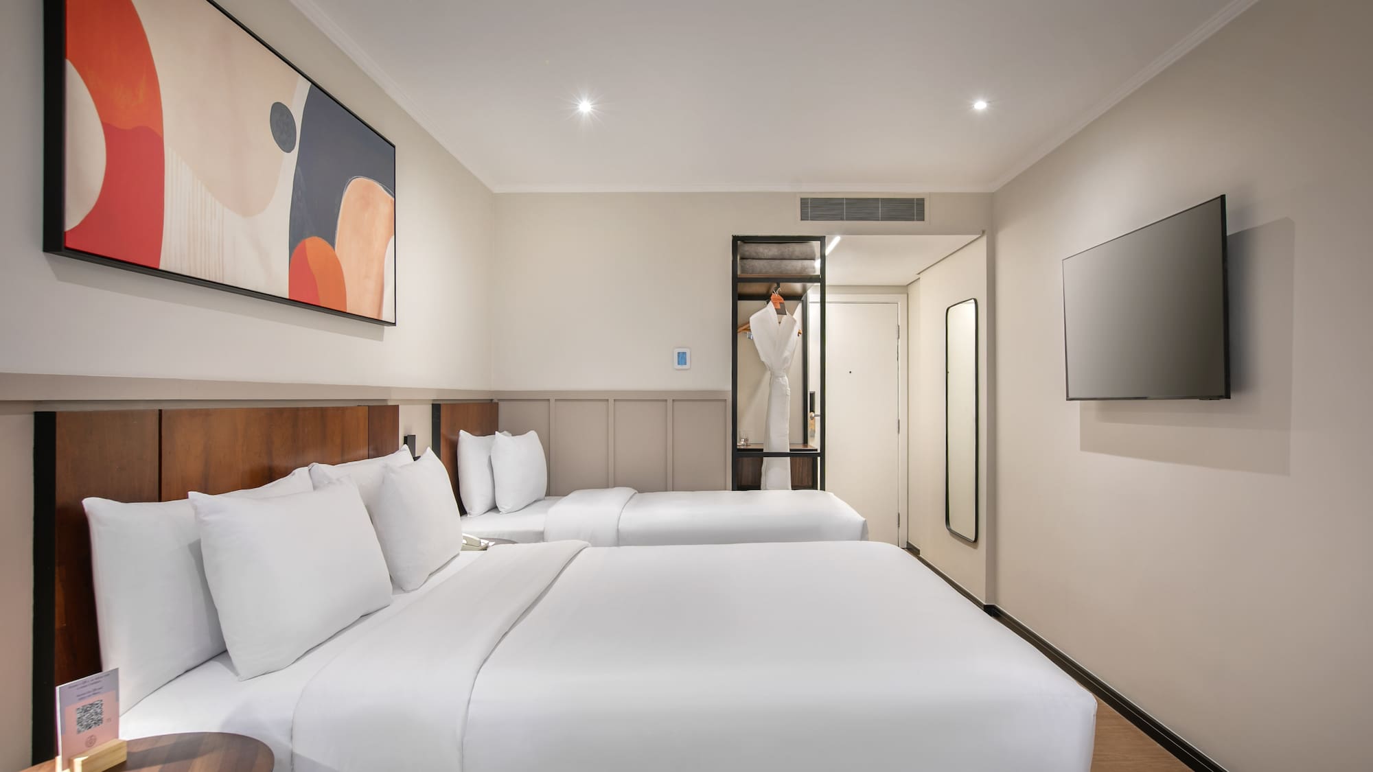 Modern hotel guest room with two beds, abstract art, TV, and open wardrobe.