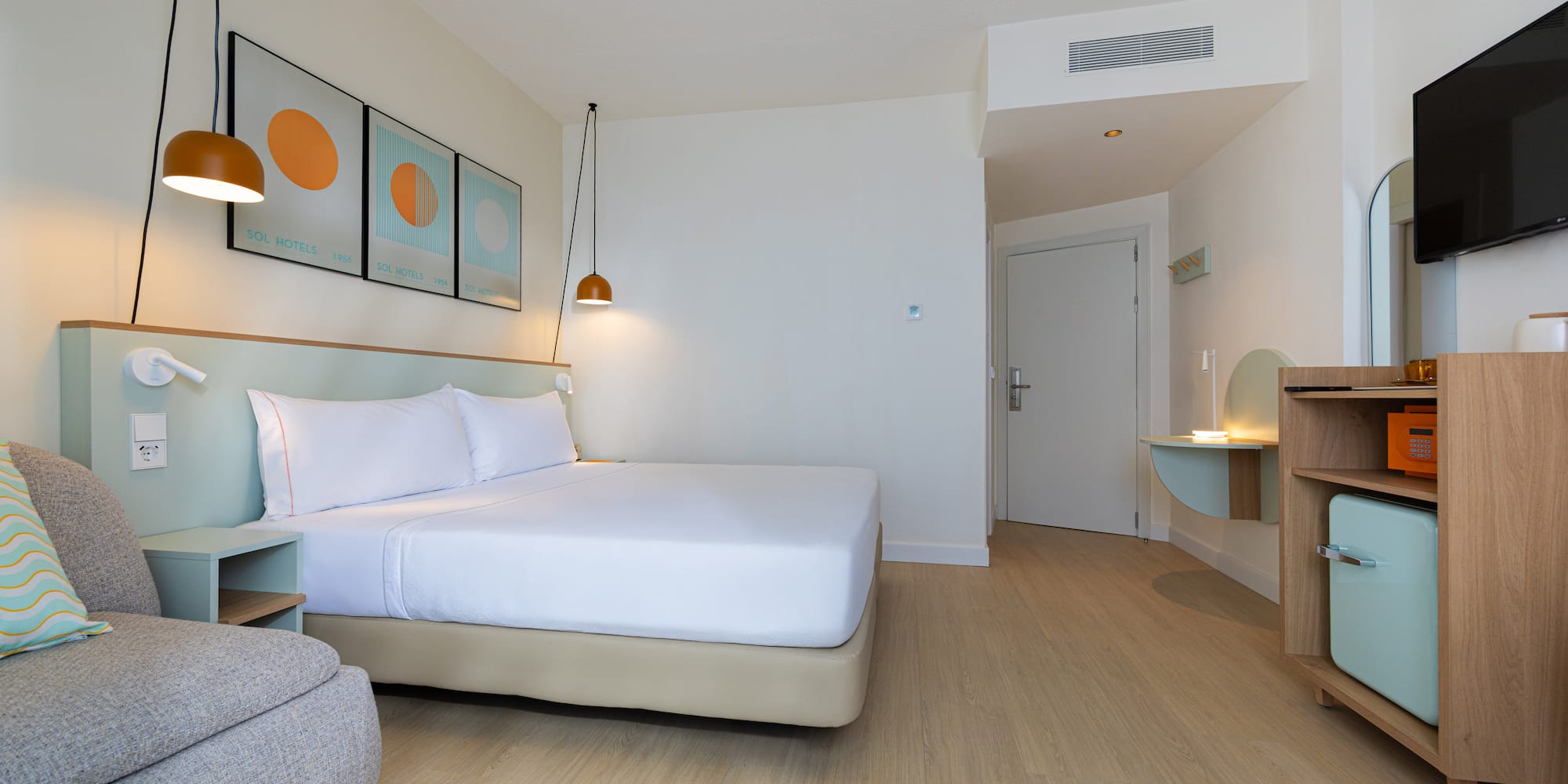 Modern hotel room with king bed, wood floor, mini-fridge, and TV.