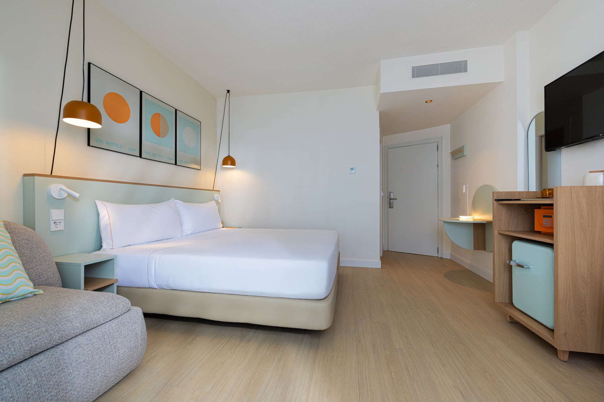 Contemporary hotel room with white bed, wooden floor, armchair, and mini-fridge.