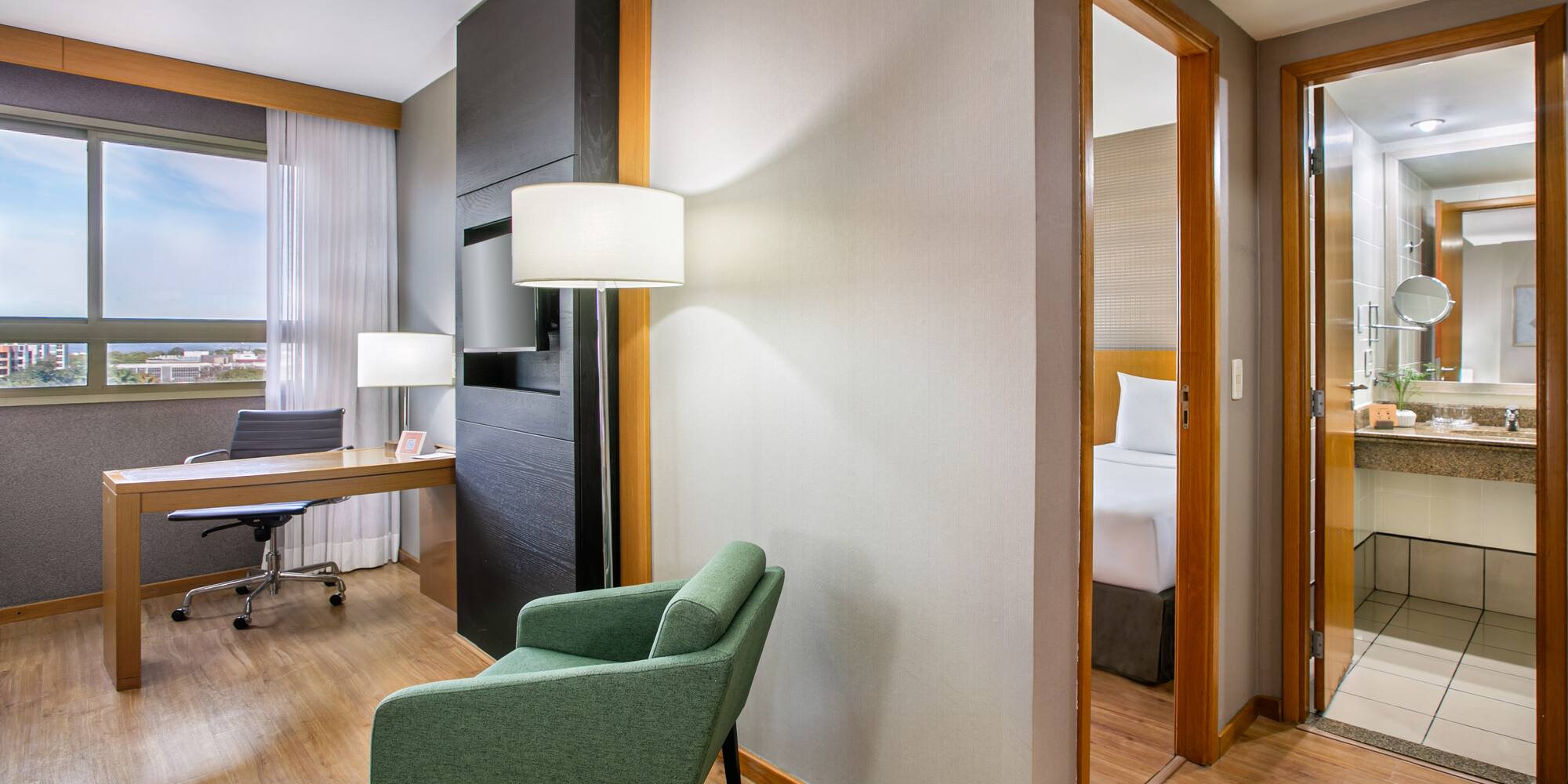 Modern hotel suite with workspace, green armchair, bedroom, and bathroom.