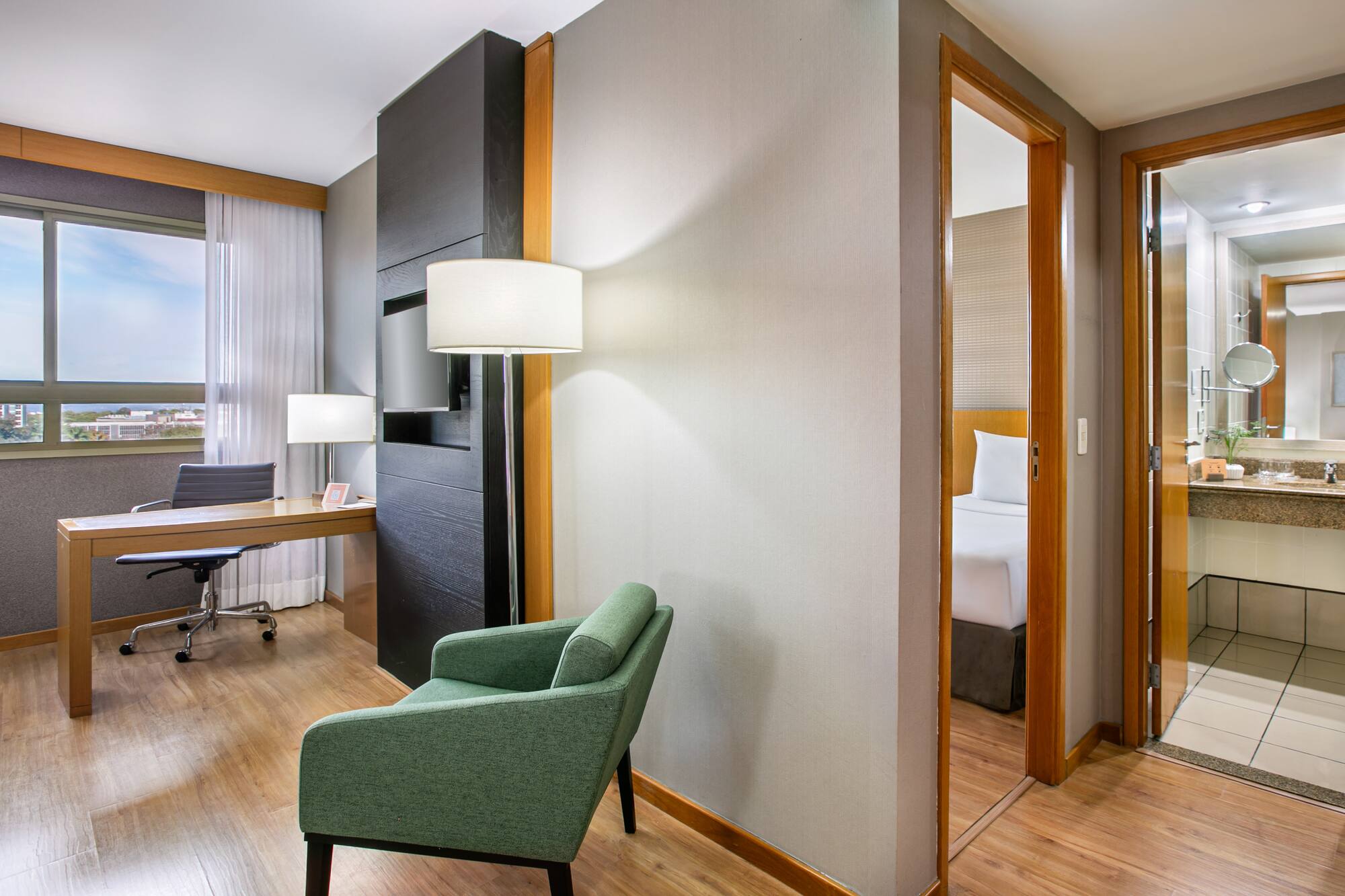 Modern hotel suite with workspace, green armchair, bedroom, and bathroom.