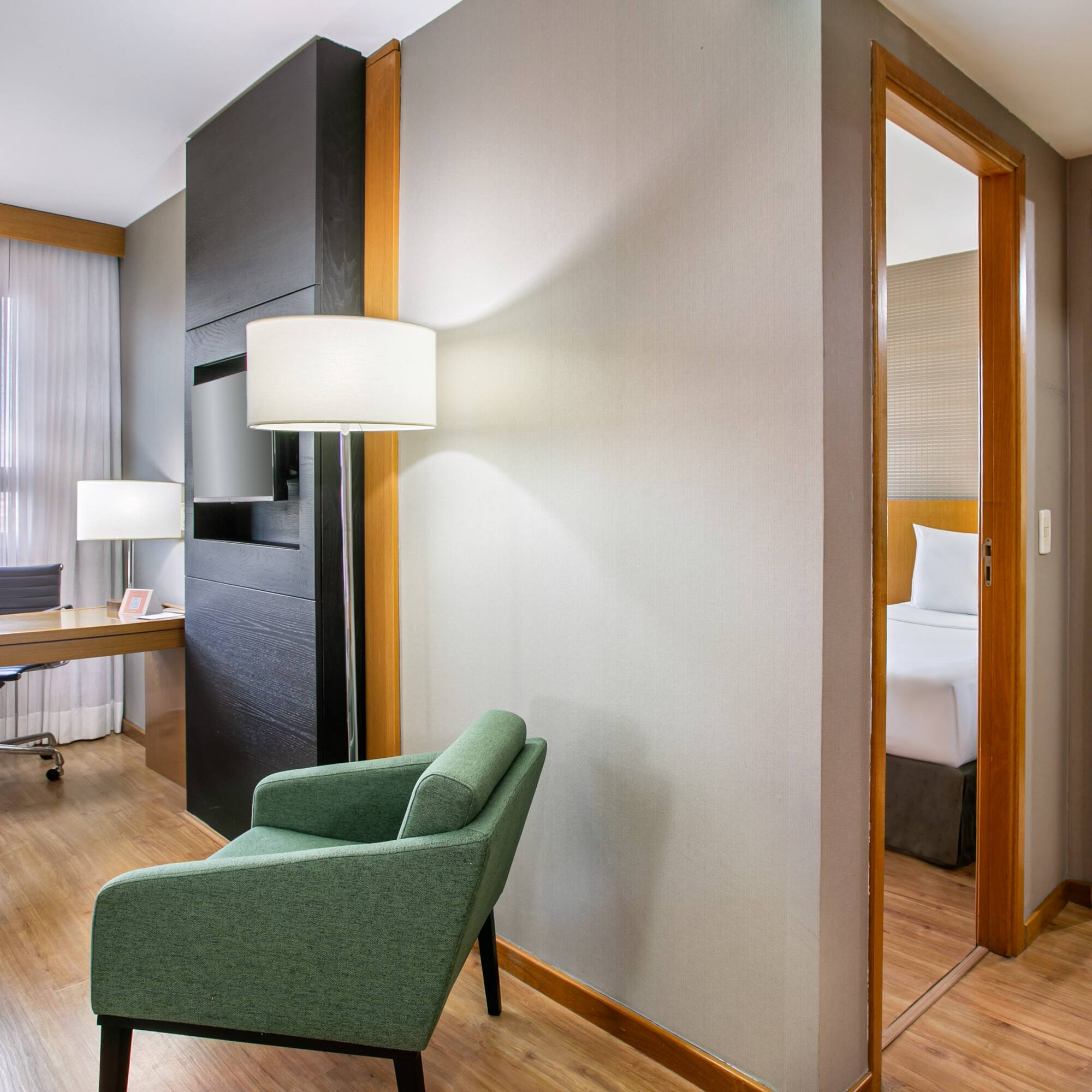 Modern hotel suite with workspace, green armchair, bedroom, and bathroom.