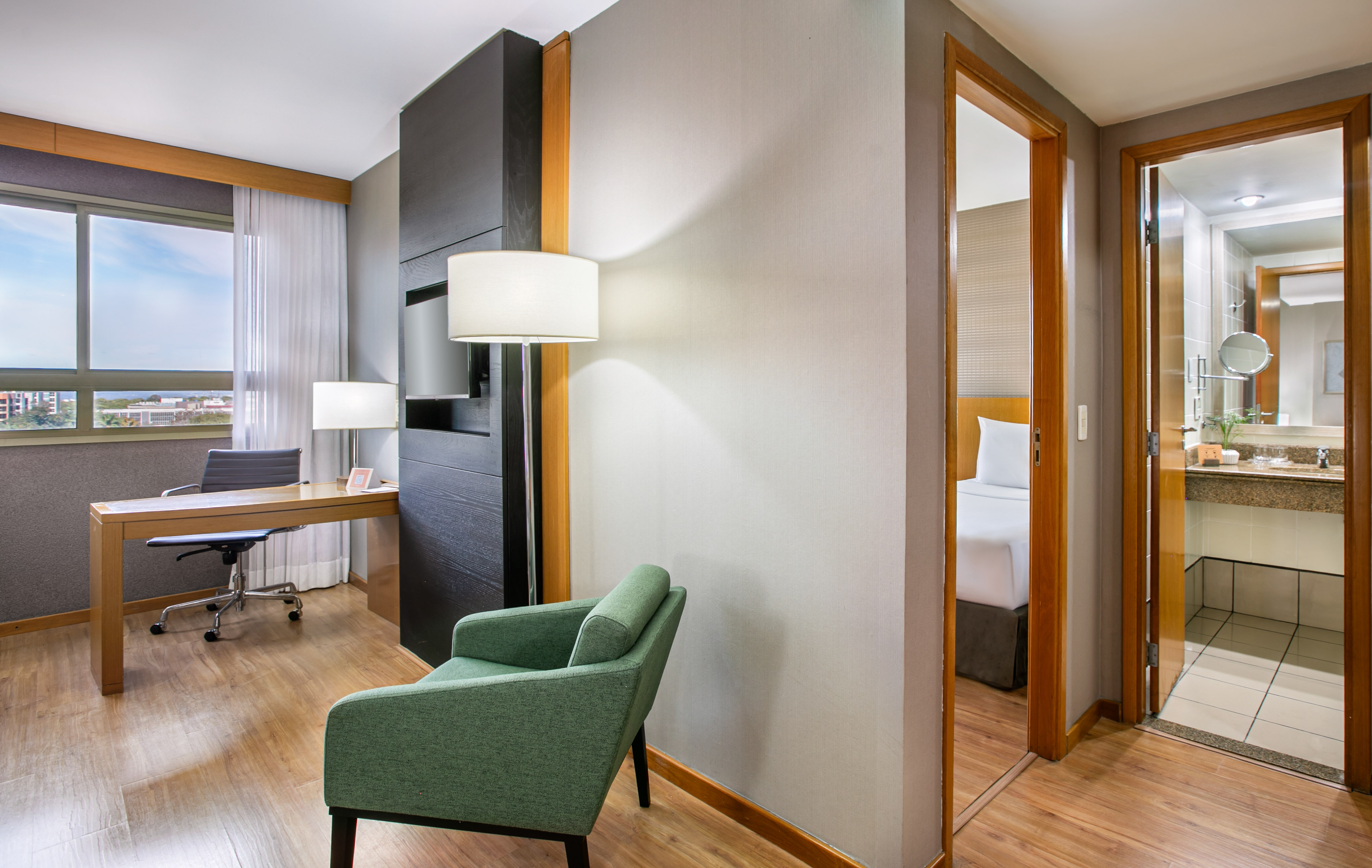 Modern hotel suite with workspace, green armchair, bedroom, and bathroom.