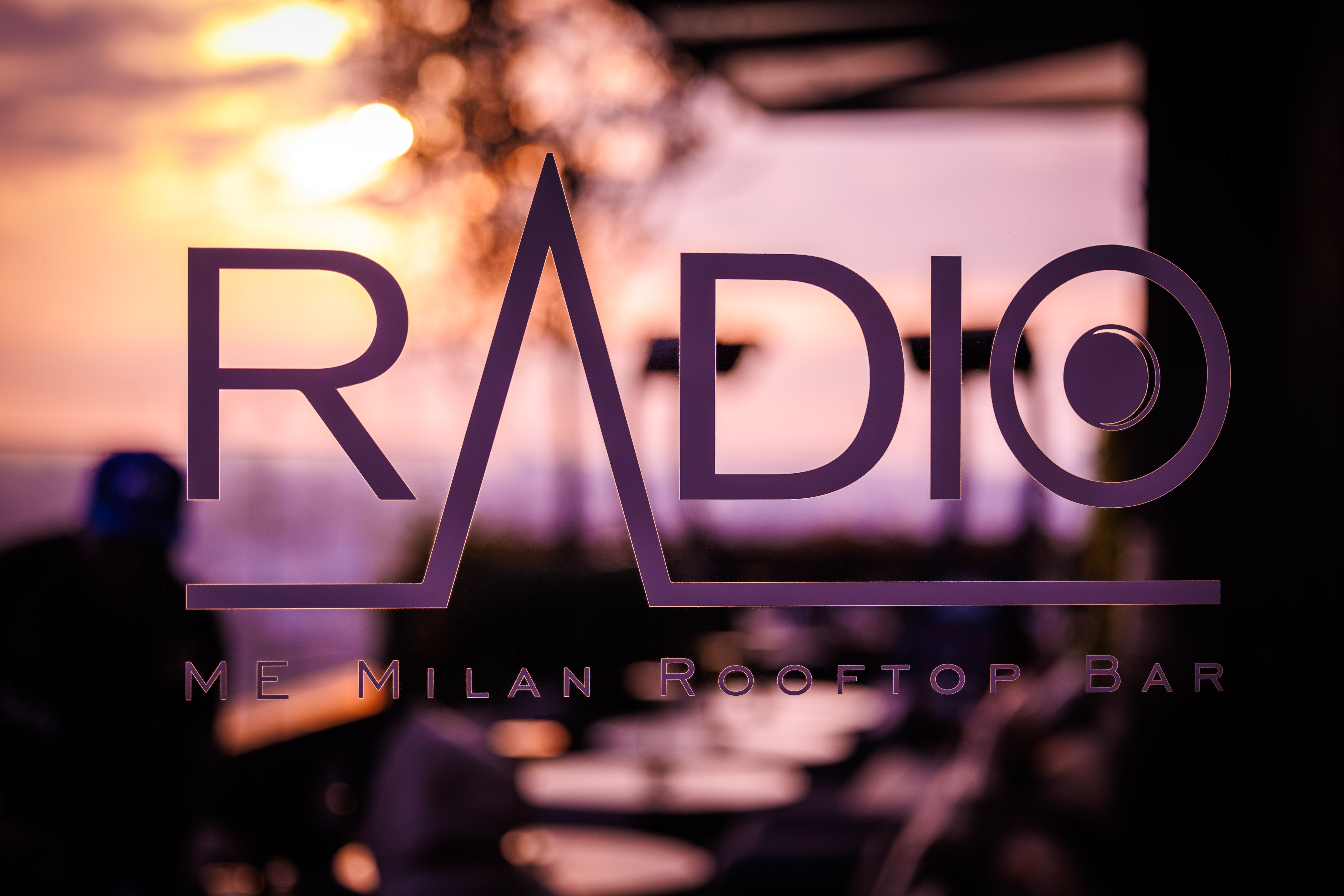 Radio ME Milan Rooftop Bar logo, purple text against blurred sunset sky.