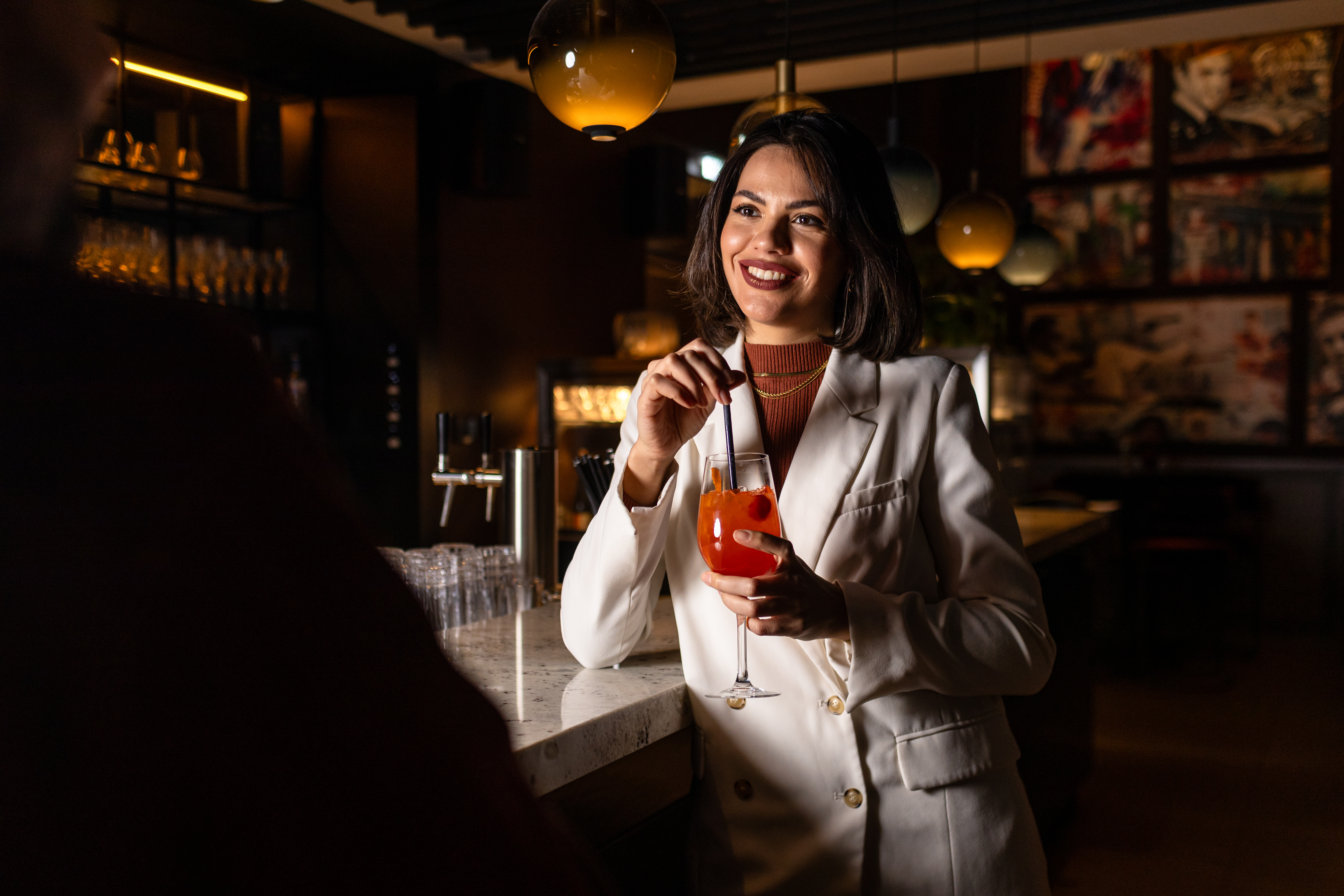 Smiling woman in white blazer holding orange cocktail at bar.