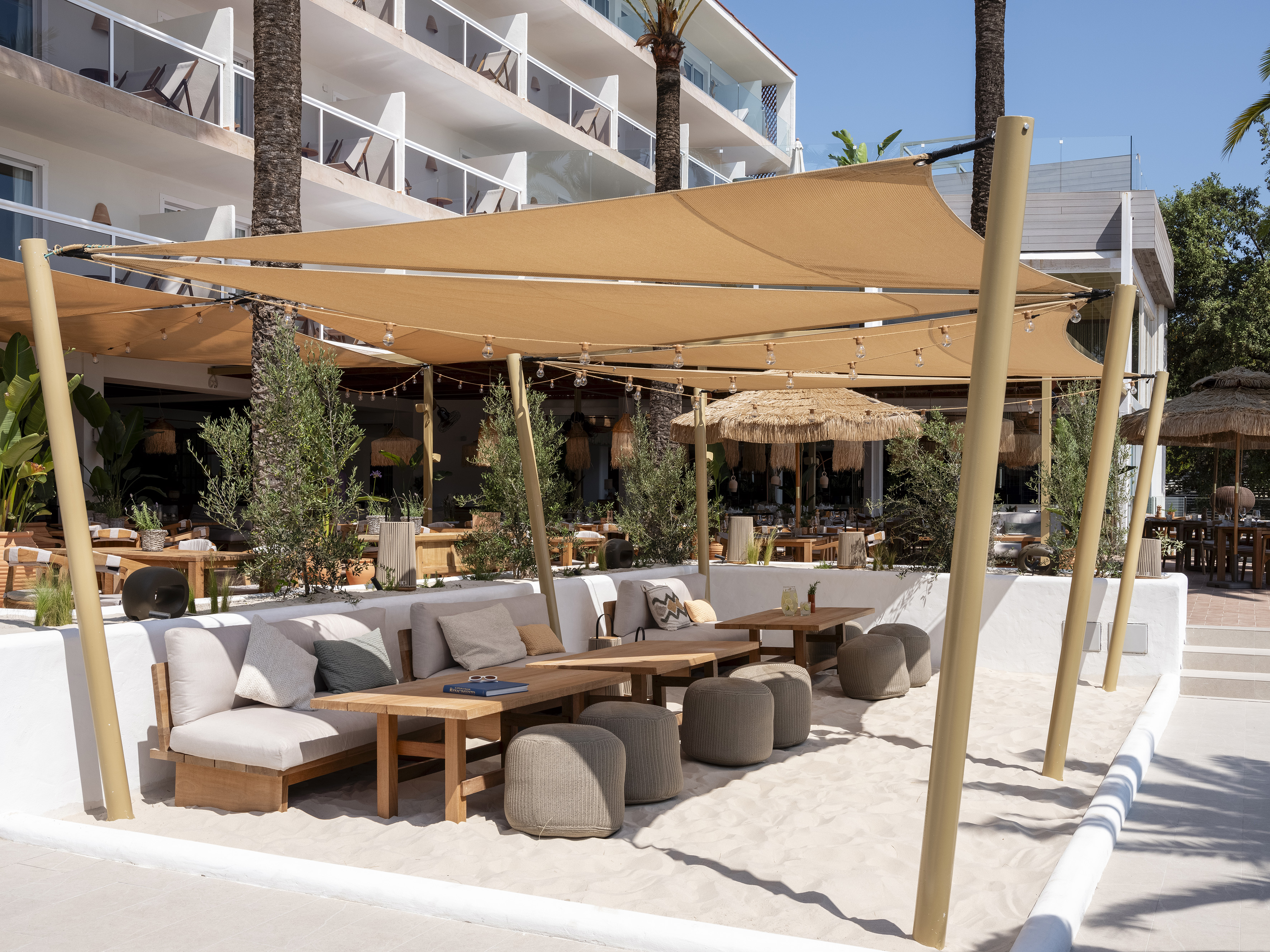 Outdoor resort dining lounge with sand floor and large beige shade canopy.