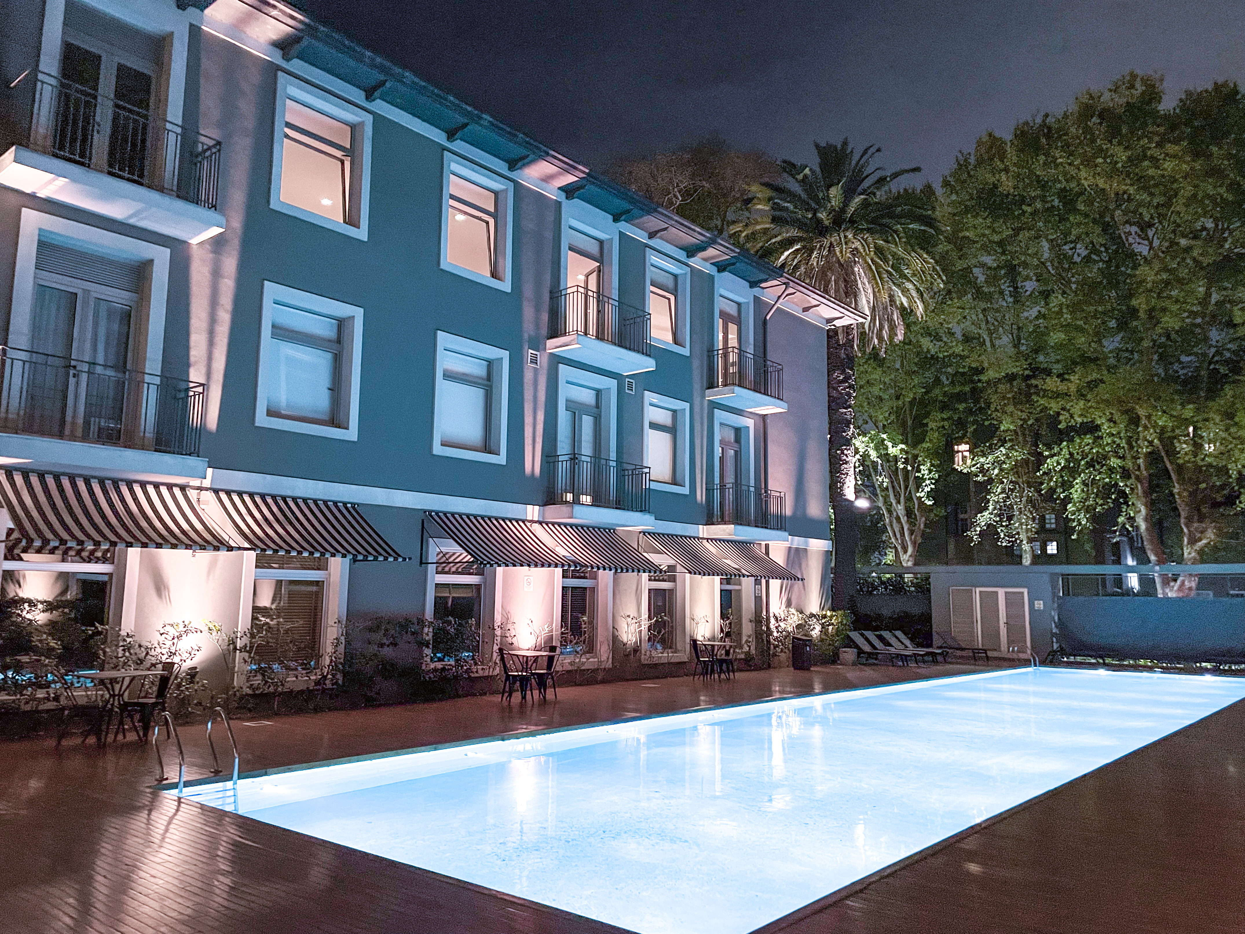 Hotel facade at night with illuminated swimming pool and striped awnings.