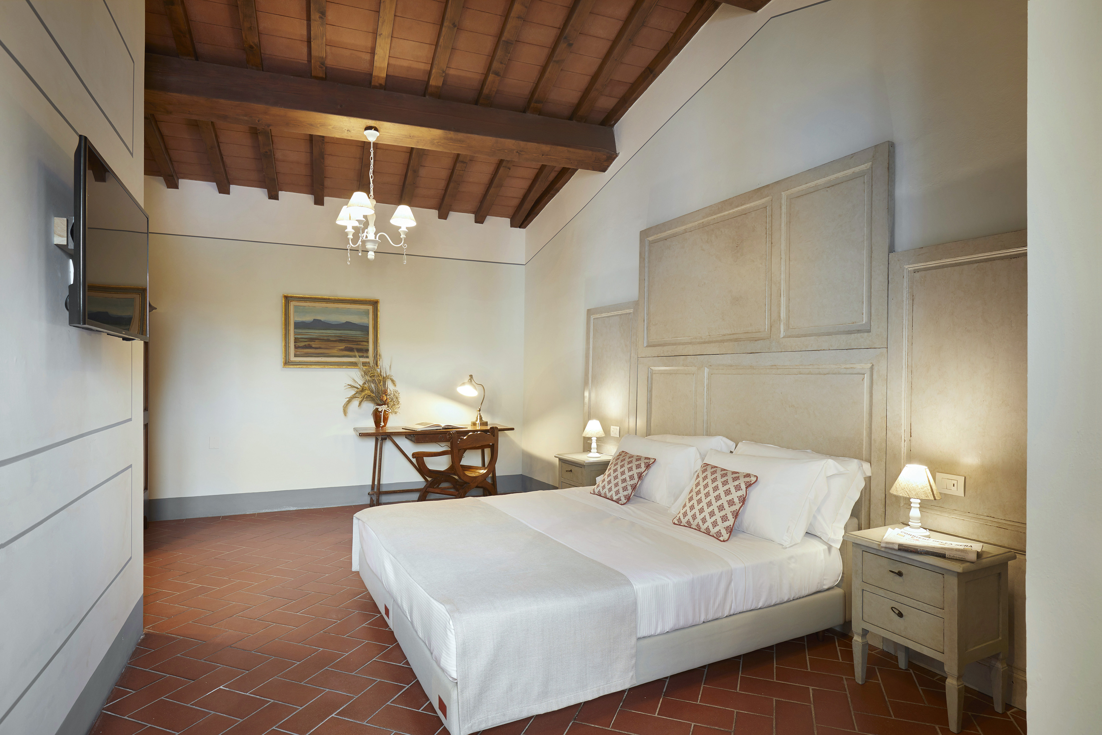 Tuscan bedroom with large bed, terracotta floor, wooden beamed ceiling, and desk.