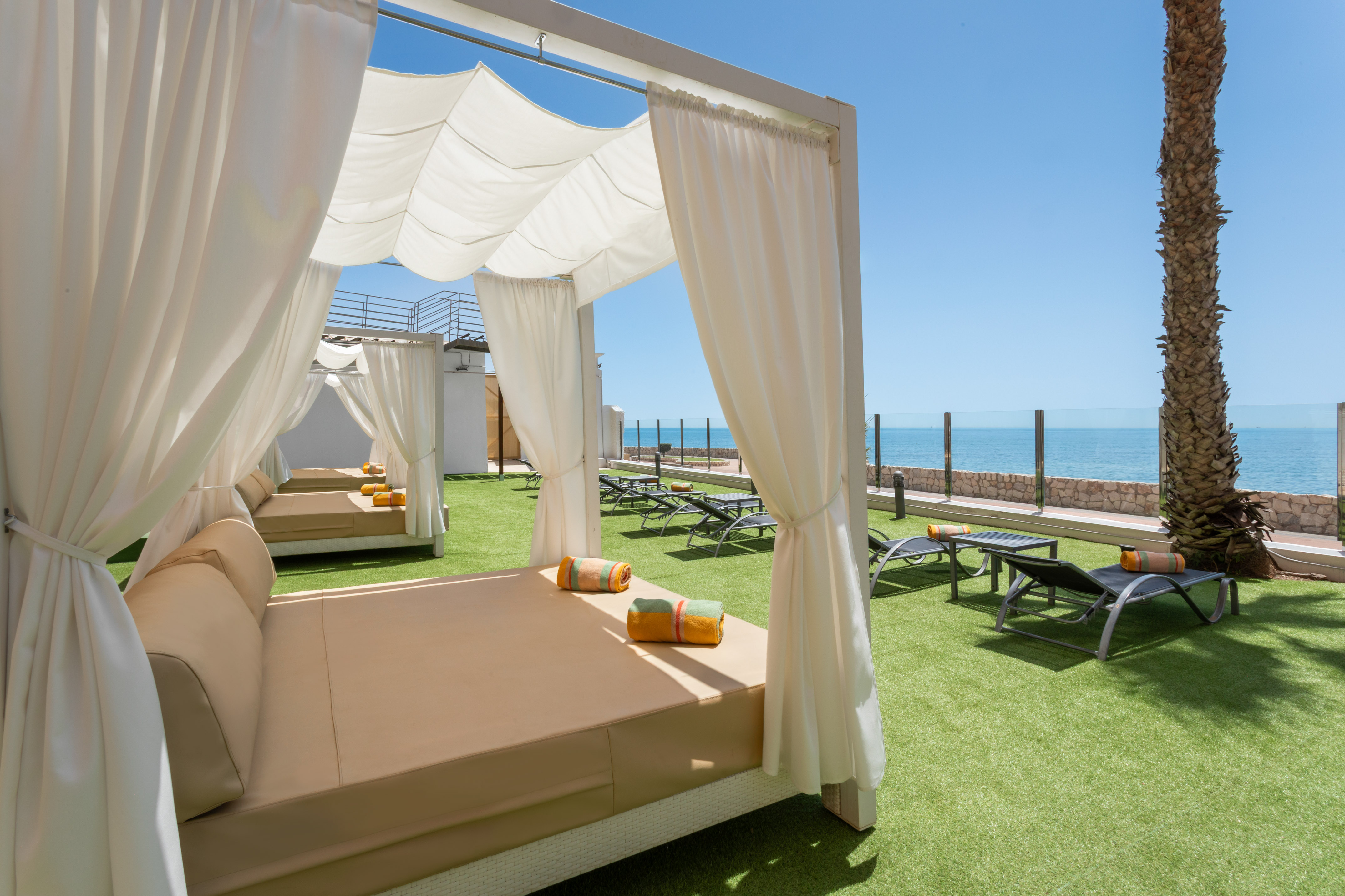 Oceanfront cabana with daybeds on artificial grass, clear sky and sea view.