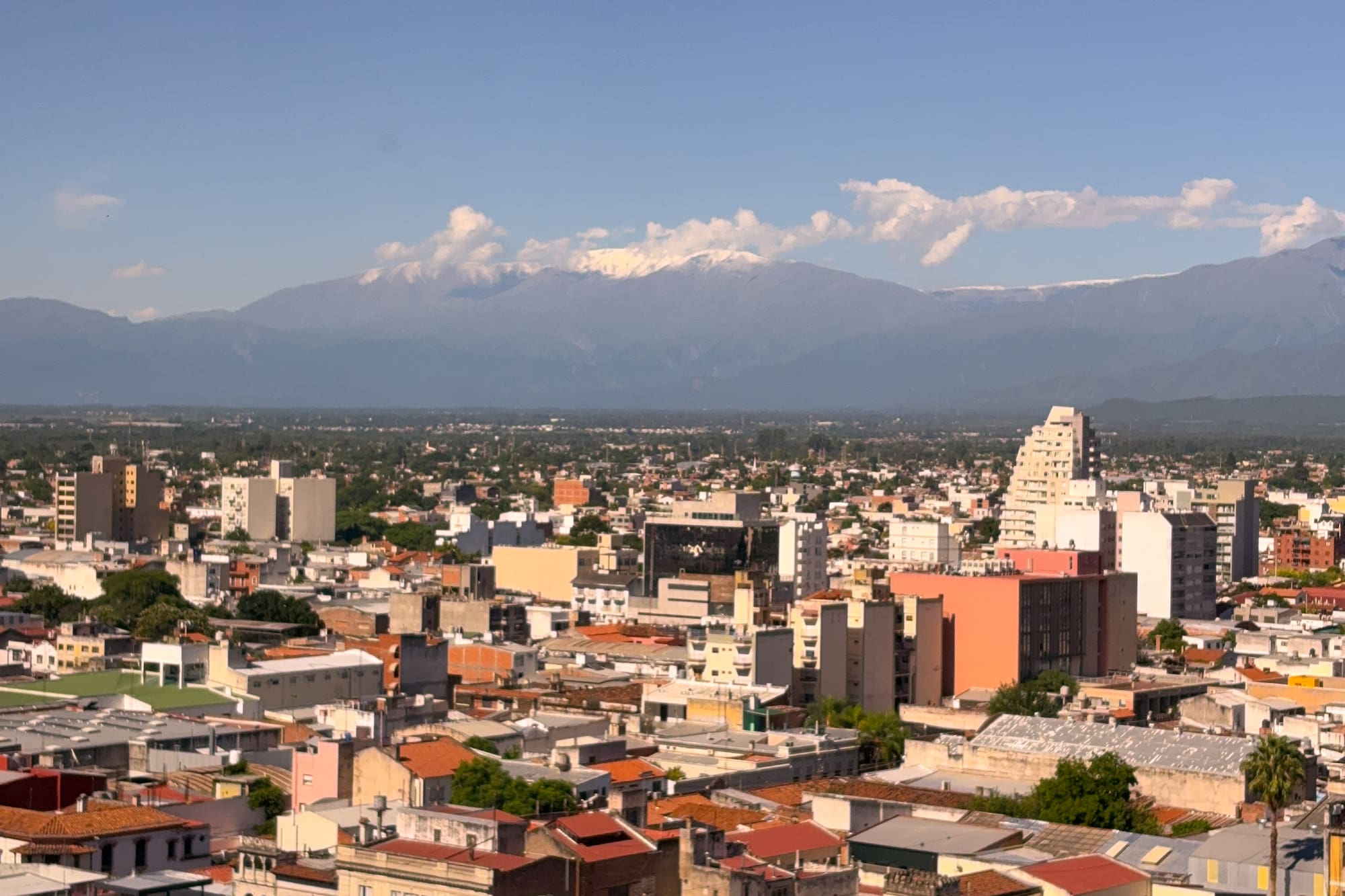 City skyline with buildings and distant snow-capped mountains under a clear blue sky.