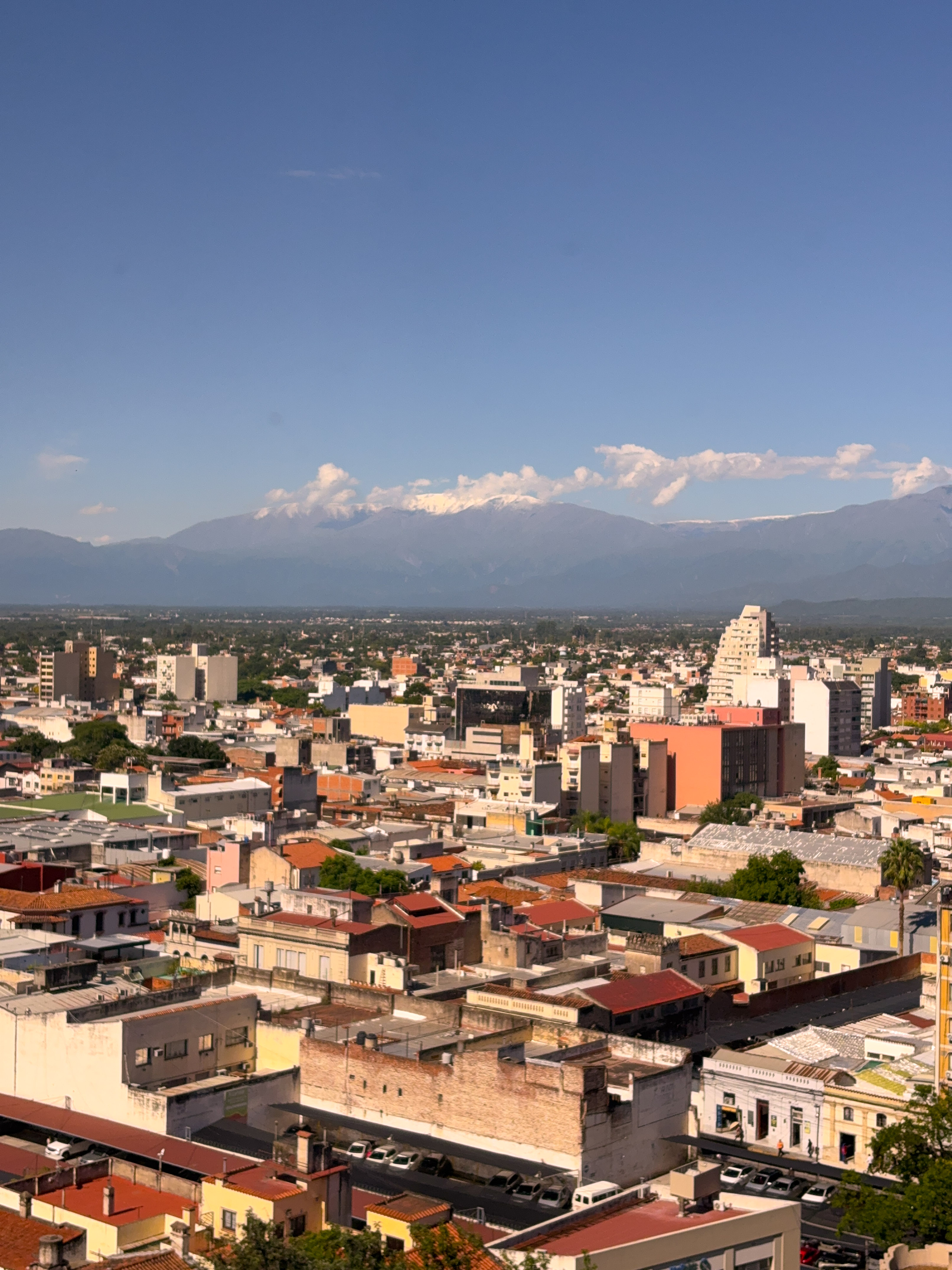 City skyline with buildings and distant snow-capped mountains under a clear blue sky.