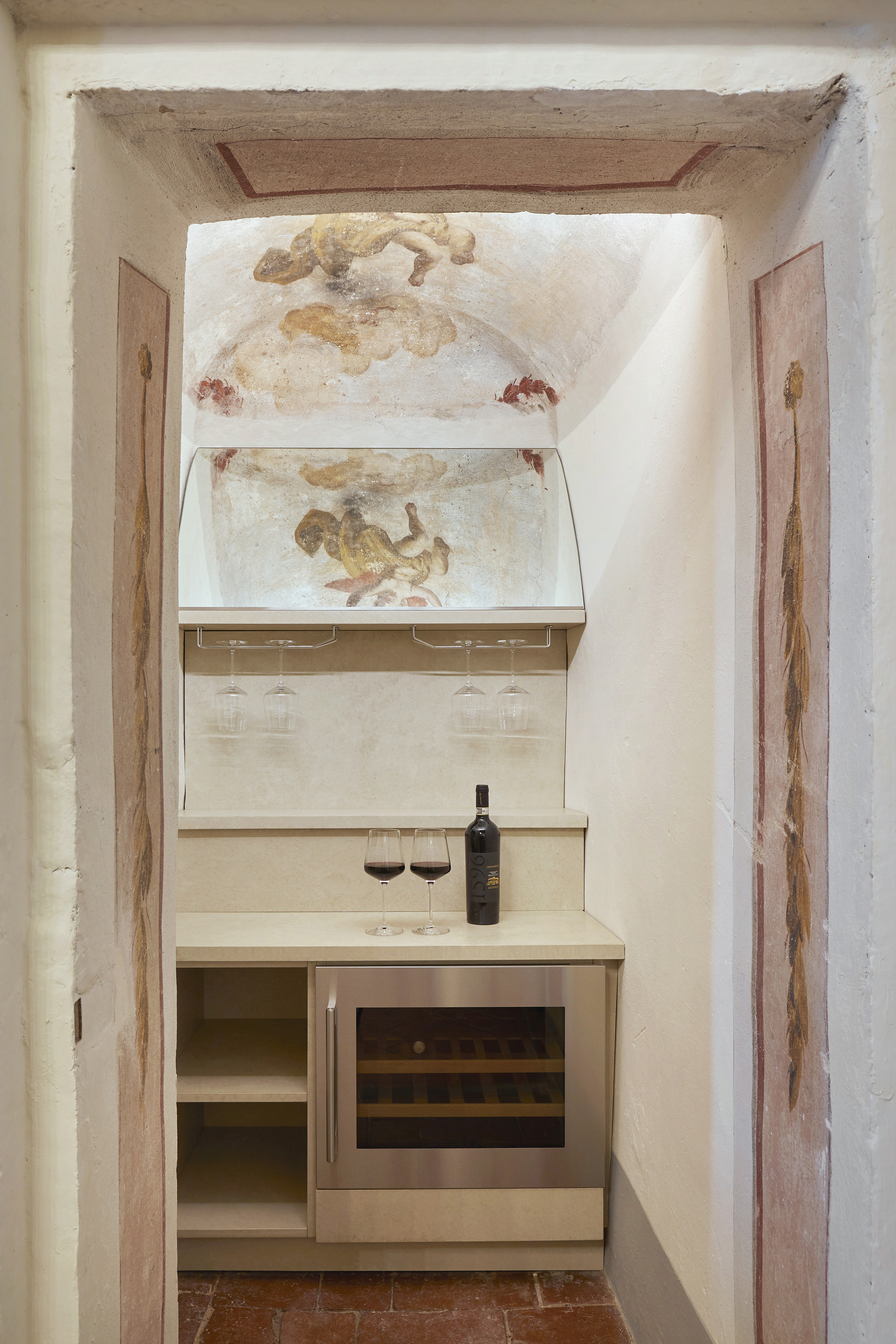 Frescoed historic wine niche with modern wine fridge, glasses, and bottle.