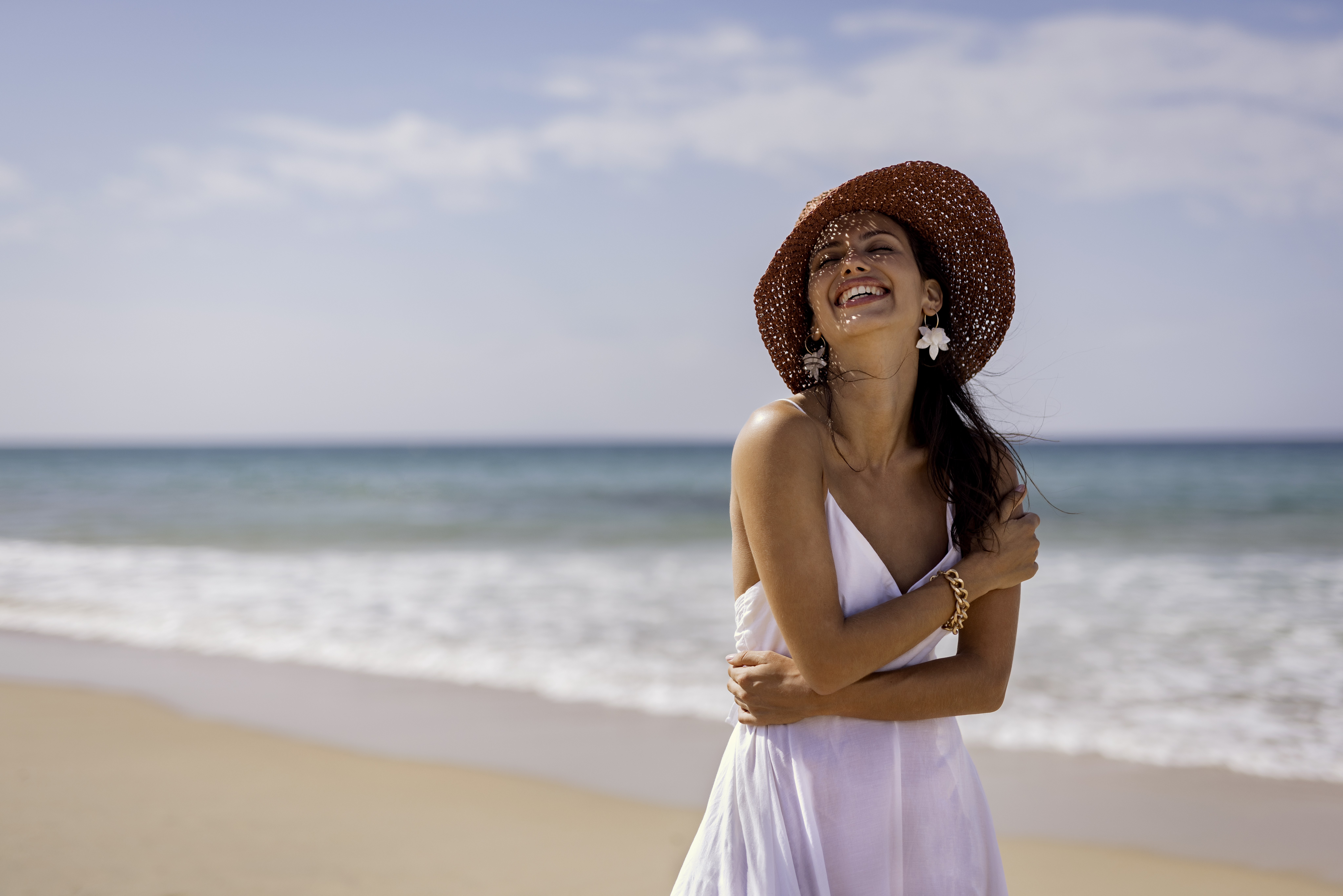 Happy woman in white dress, straw hat, laughing on sandy beach with ocean.