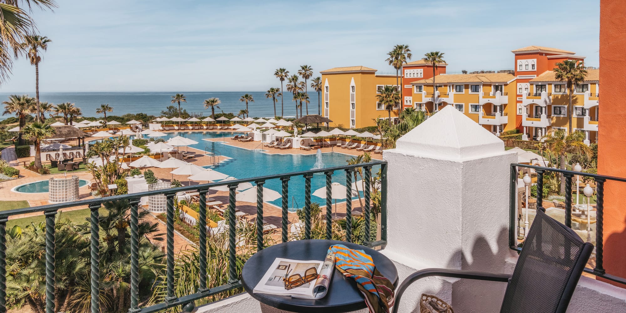Balcony view of a colorful luxury resort with pools, palm trees, and ocean.