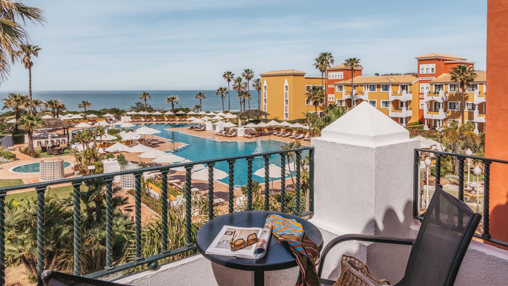 Balcony view of a colorful luxury resort with pools, palm trees, and ocean.