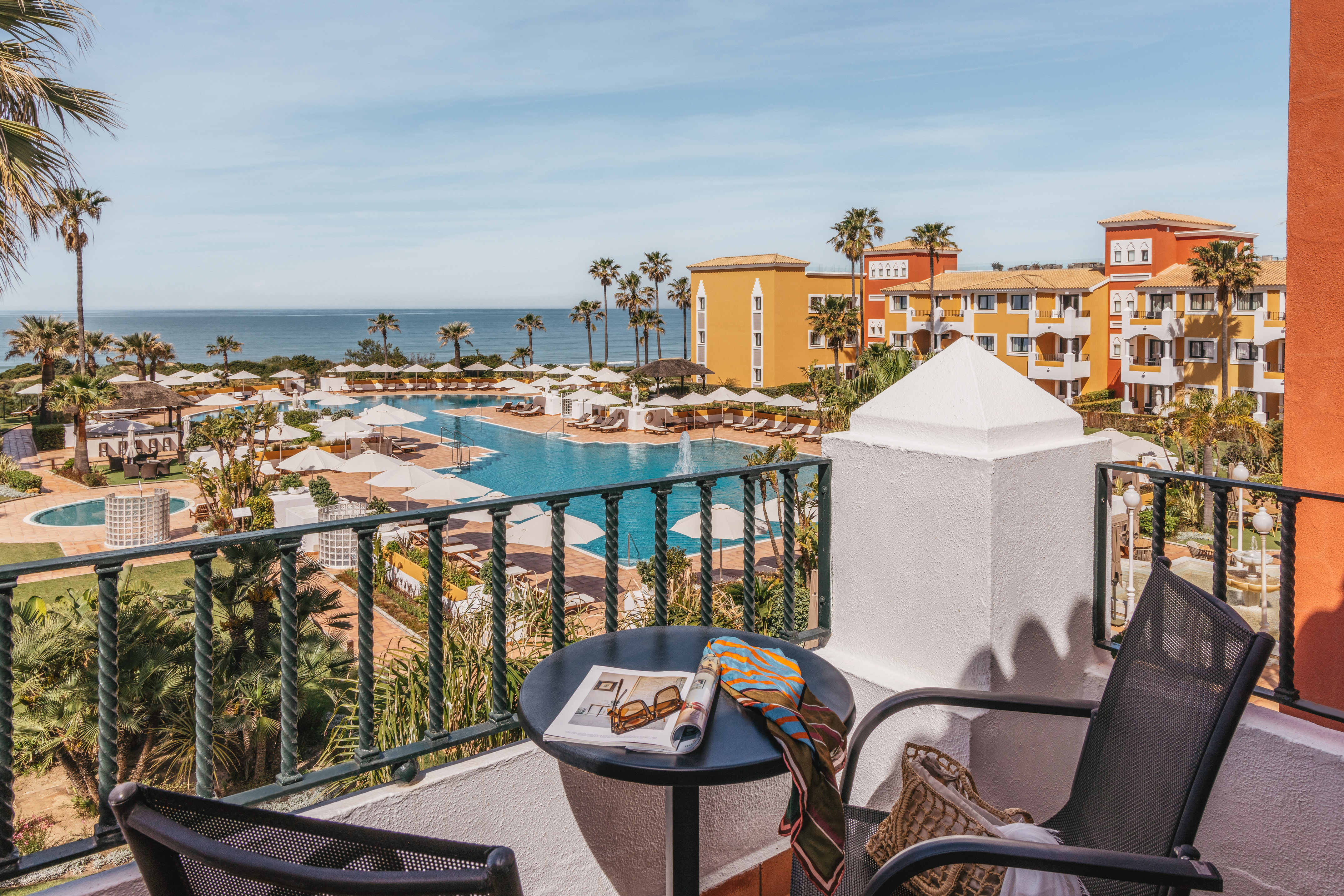Balcony view of a colorful luxury resort with pools, palm trees, and ocean.