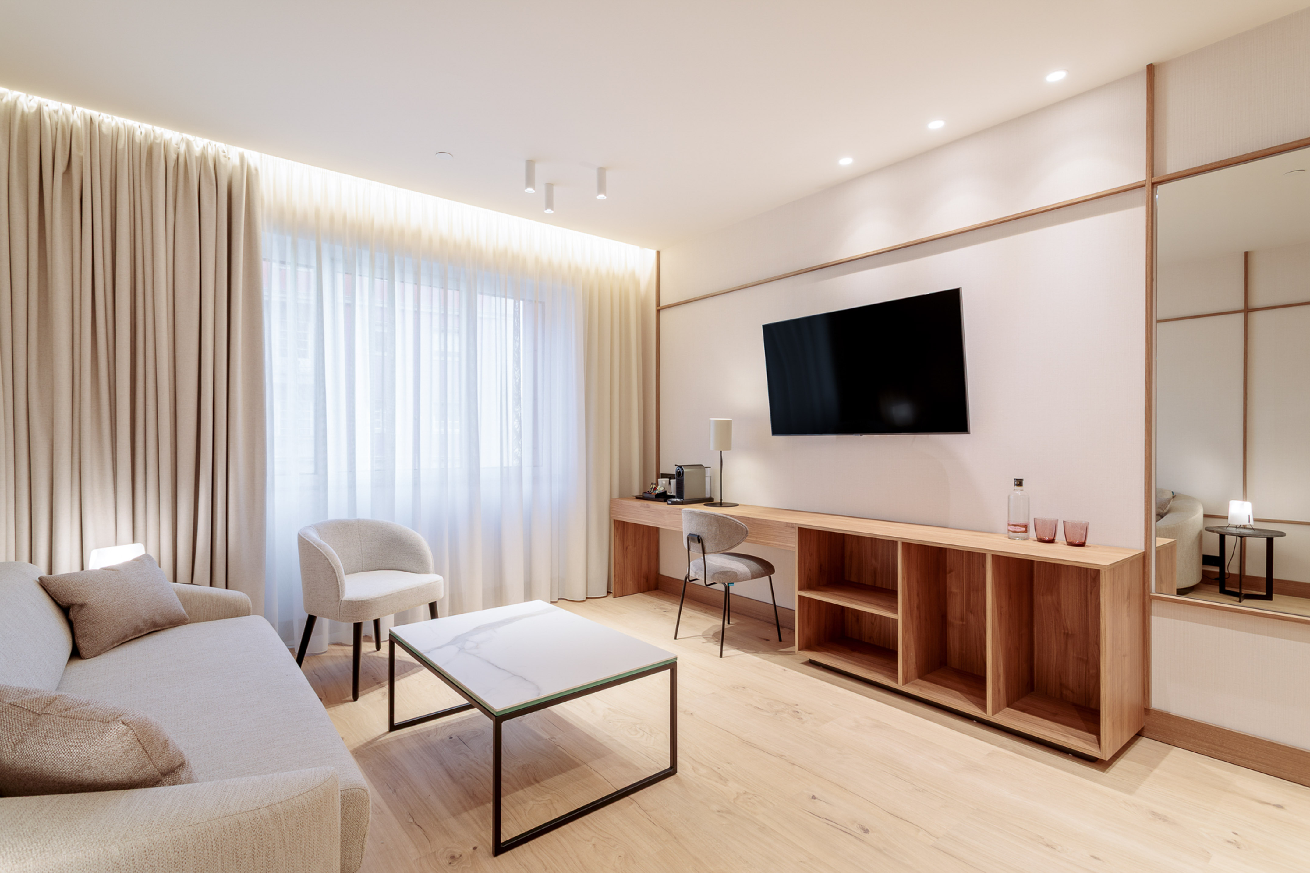 Modern hotel suite lounge with light sofa, marble table, TV, and wood desk.