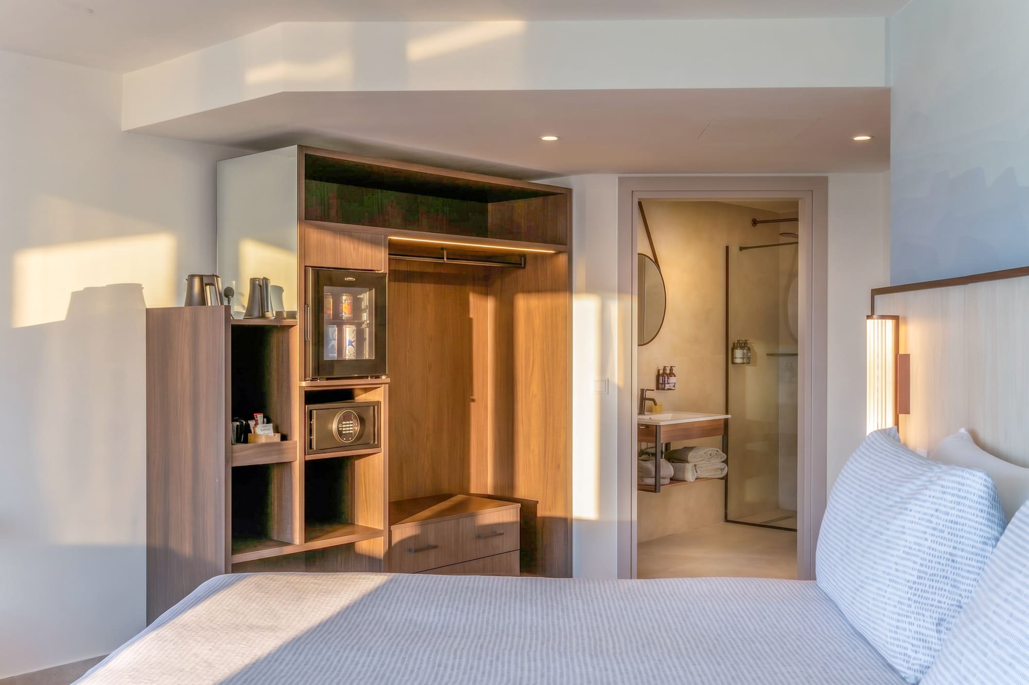 Modern hotel bedroom with bed, integrated closet, mini-bar, and bathroom.