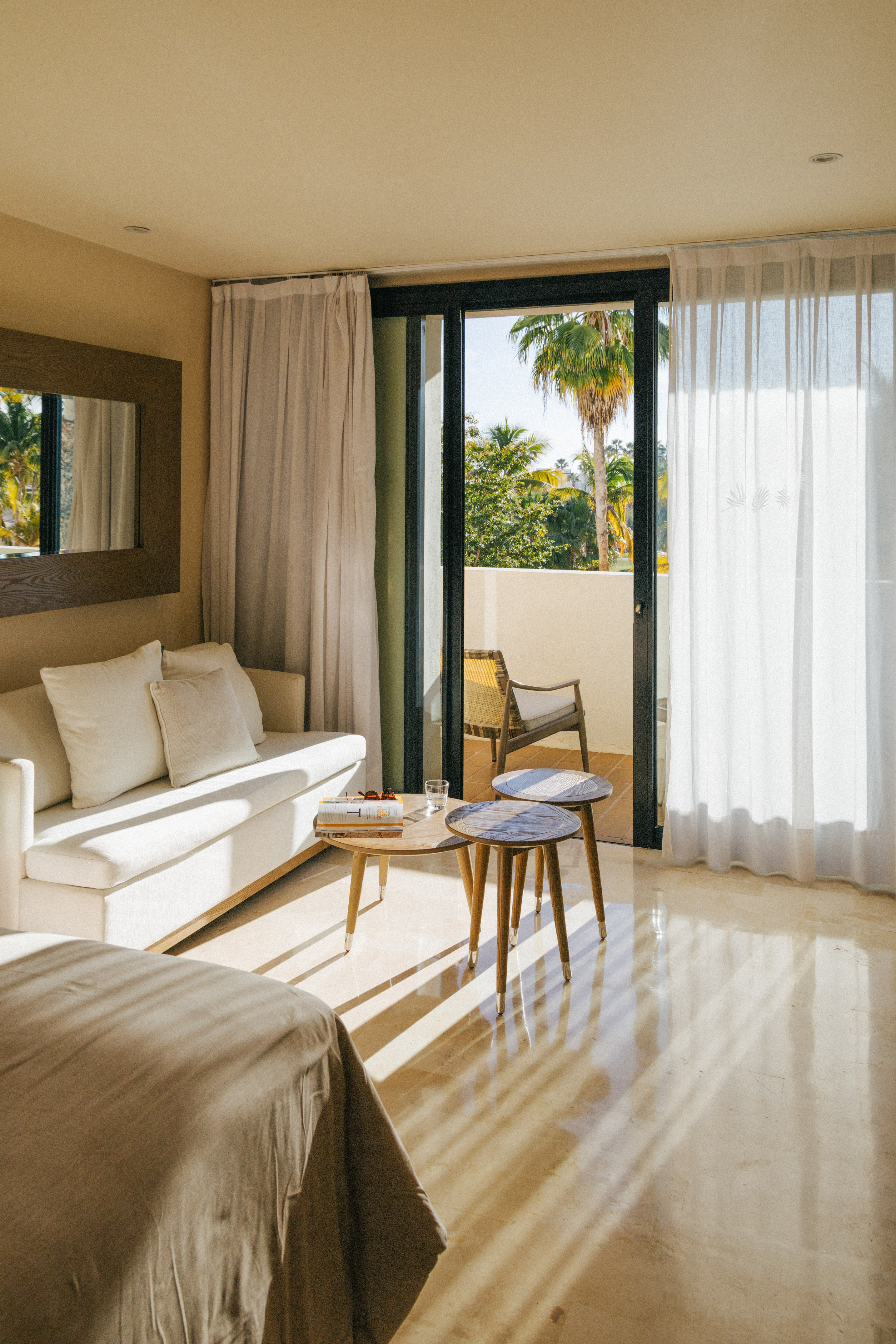 Bright hotel room with modern sofa, tables, and tropical balcony view.