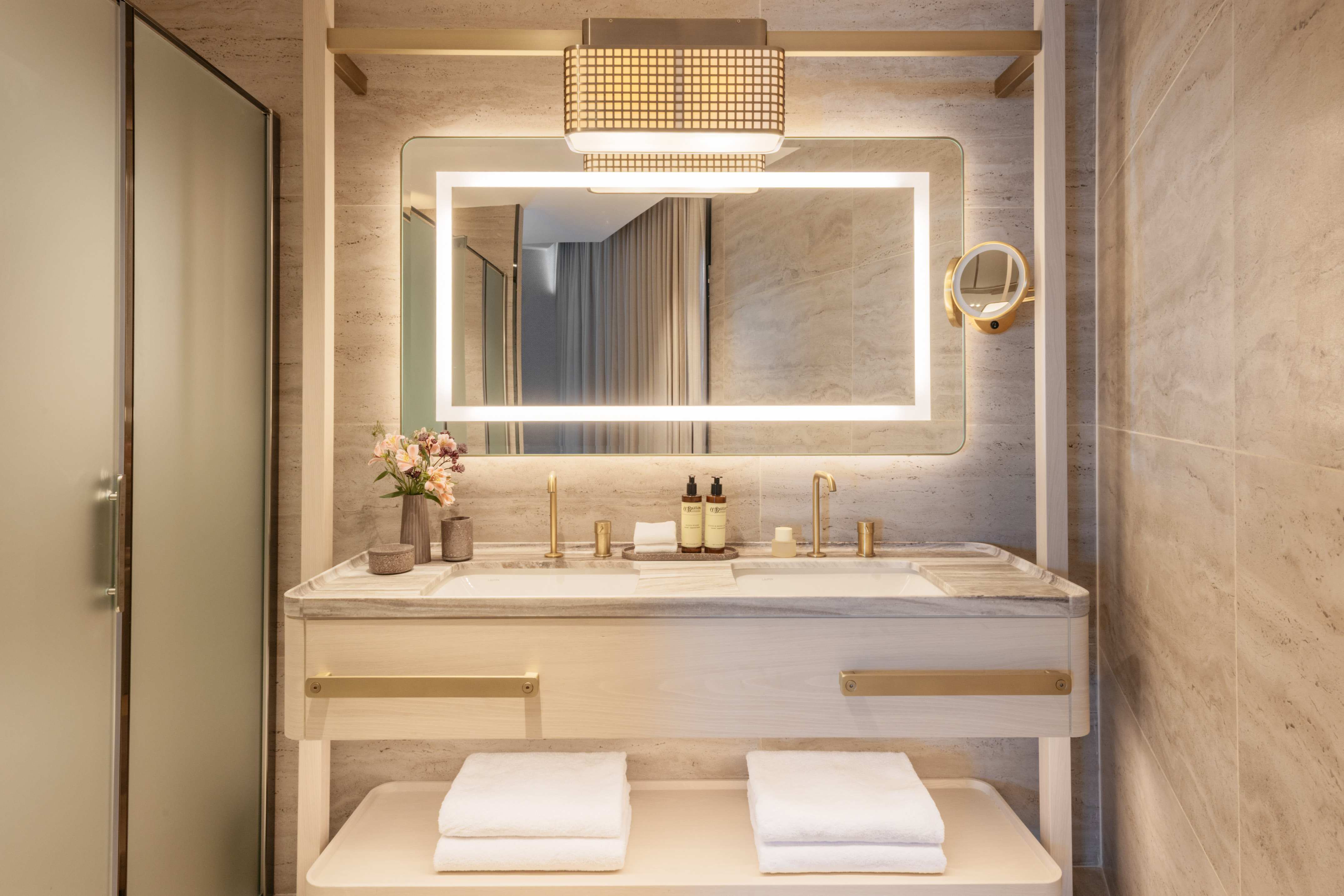 Modern hotel bathroom with double vanity, backlit mirror, and gold fixtures.