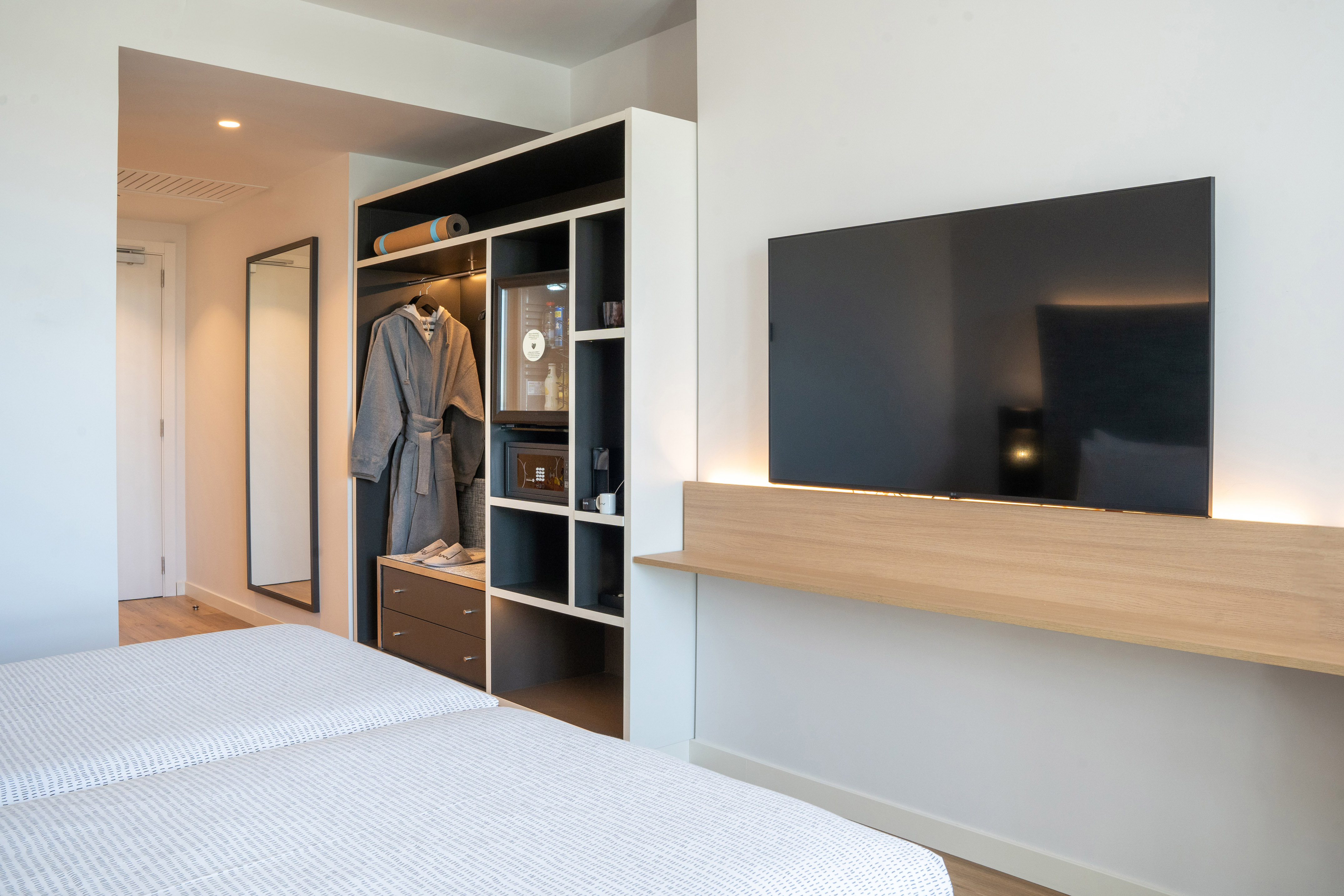 Modern hotel room: twin beds, wall TV, wardrobe, minibar, bathrobe.