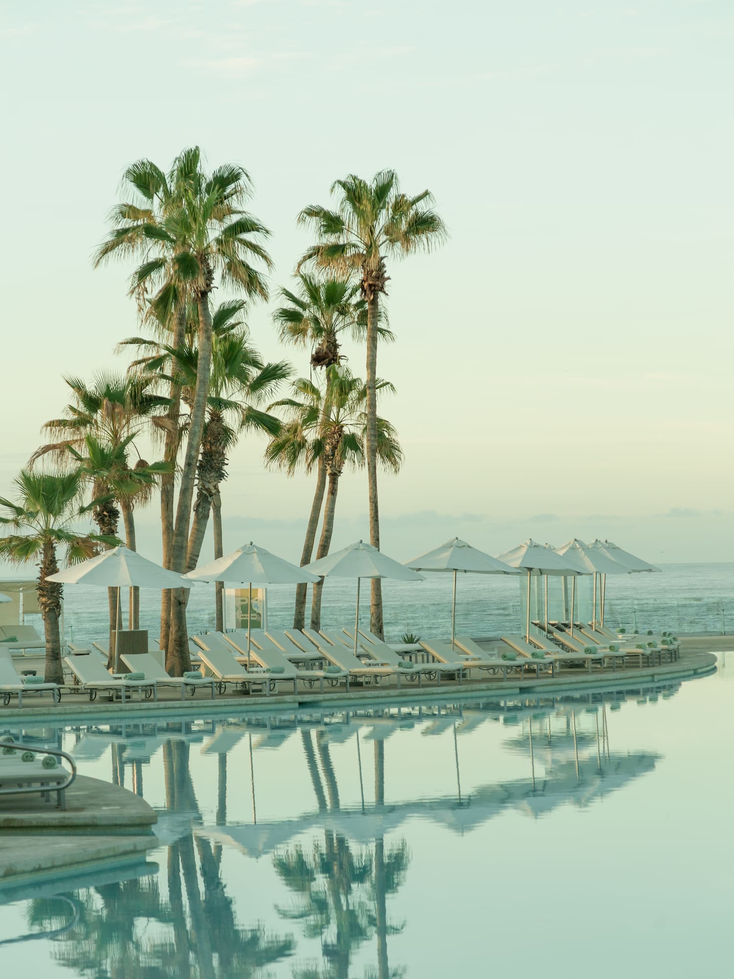 Resort pool reflecting palm trees, with sun loungers, umbrellas, and ocean.