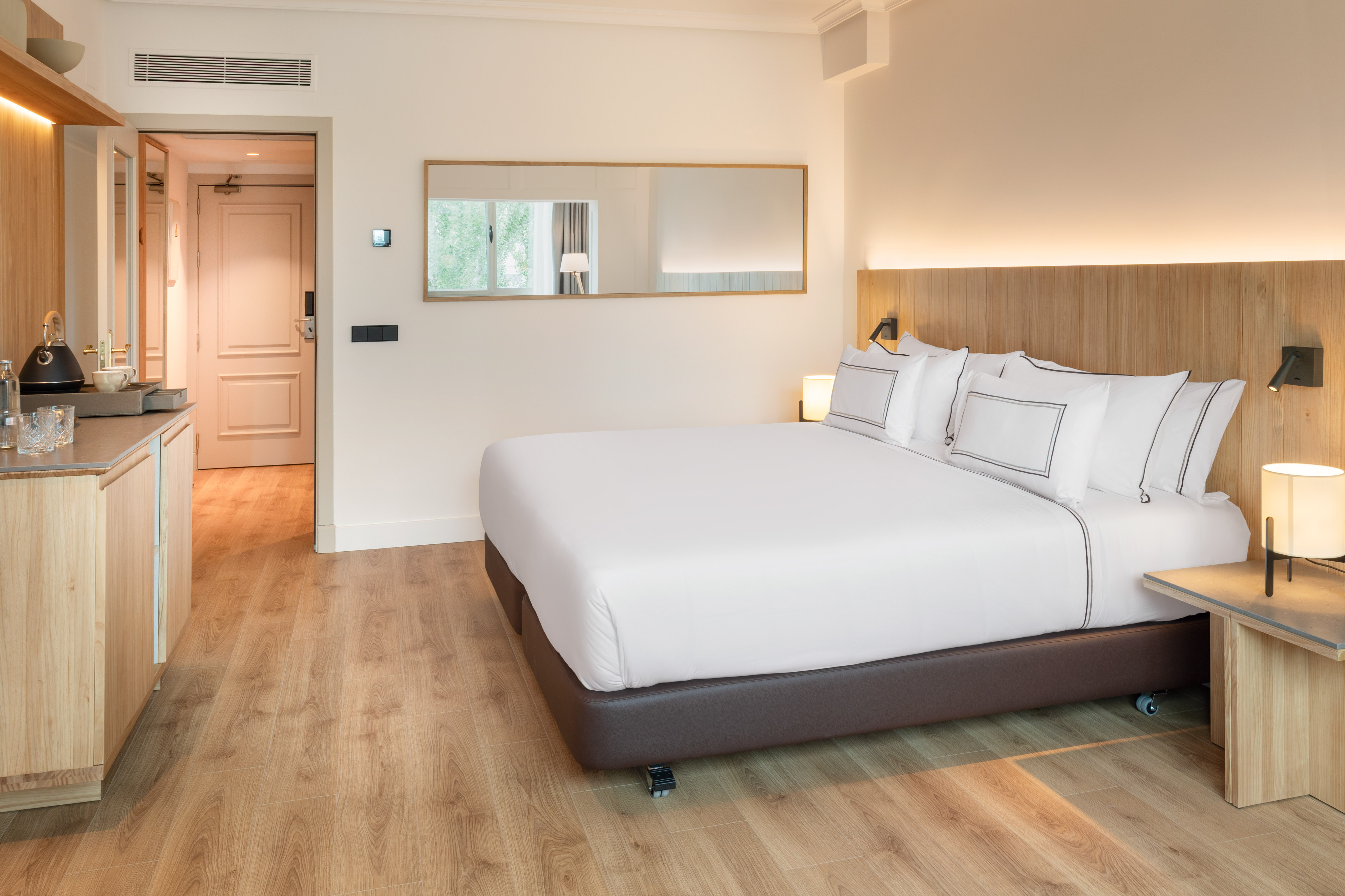 Modern hotel room with queen bed, wooden headboard, mini-bar, and light wood flooring.