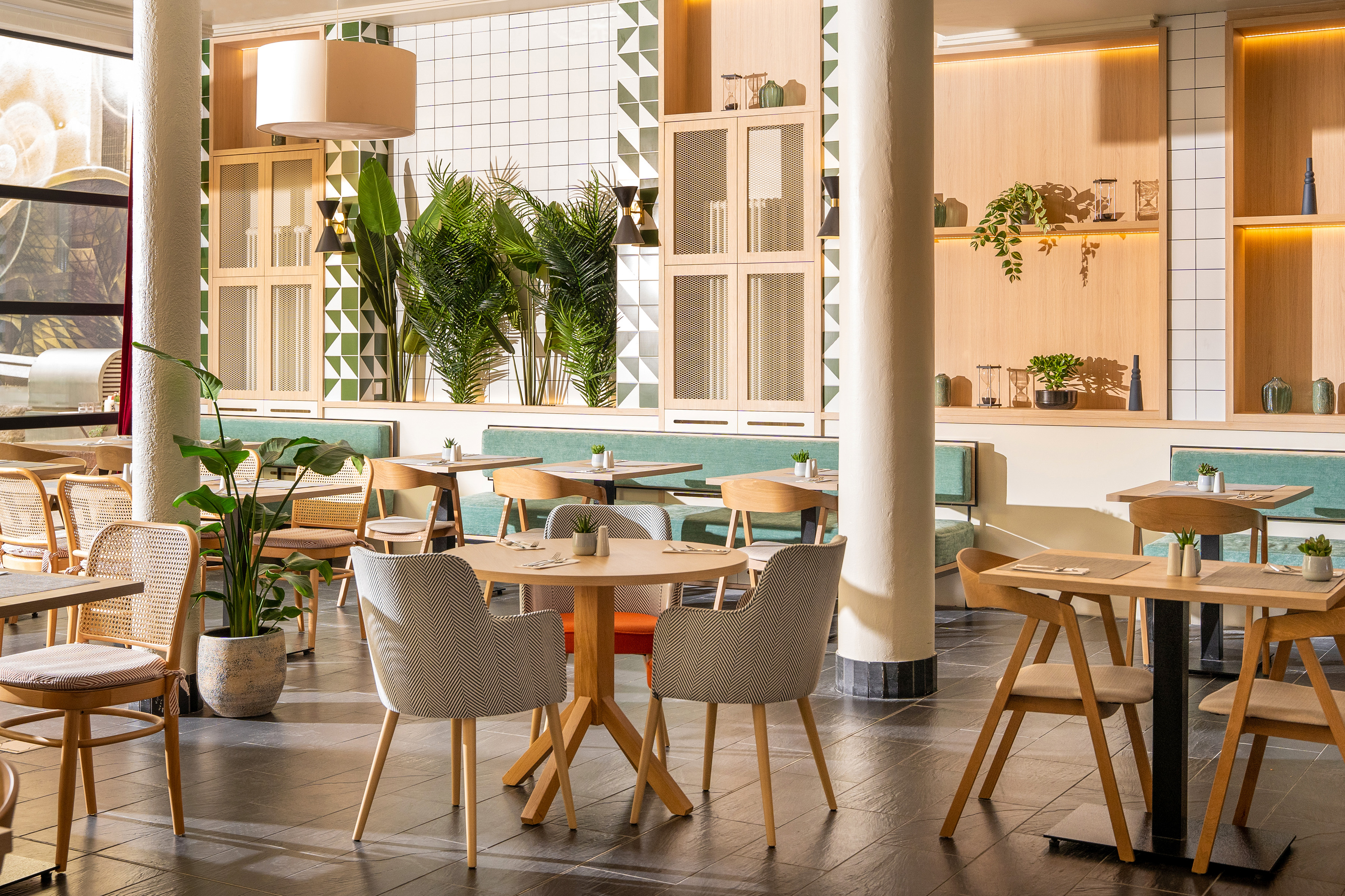 Modern cafe interior with wooden tables, chairs, lush plants, and patterned tiled walls.