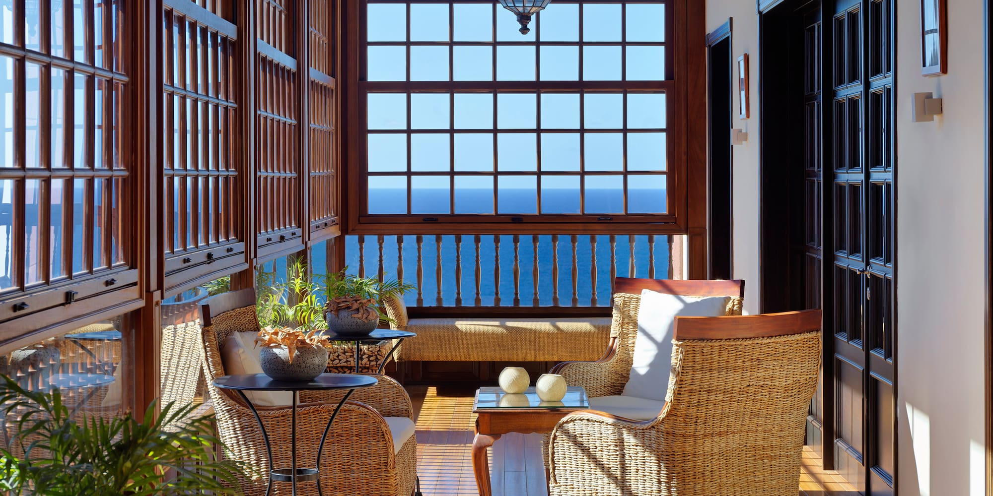 Sunny wooden sunroom with wicker chairs, sea view windows, and patterned shadows.