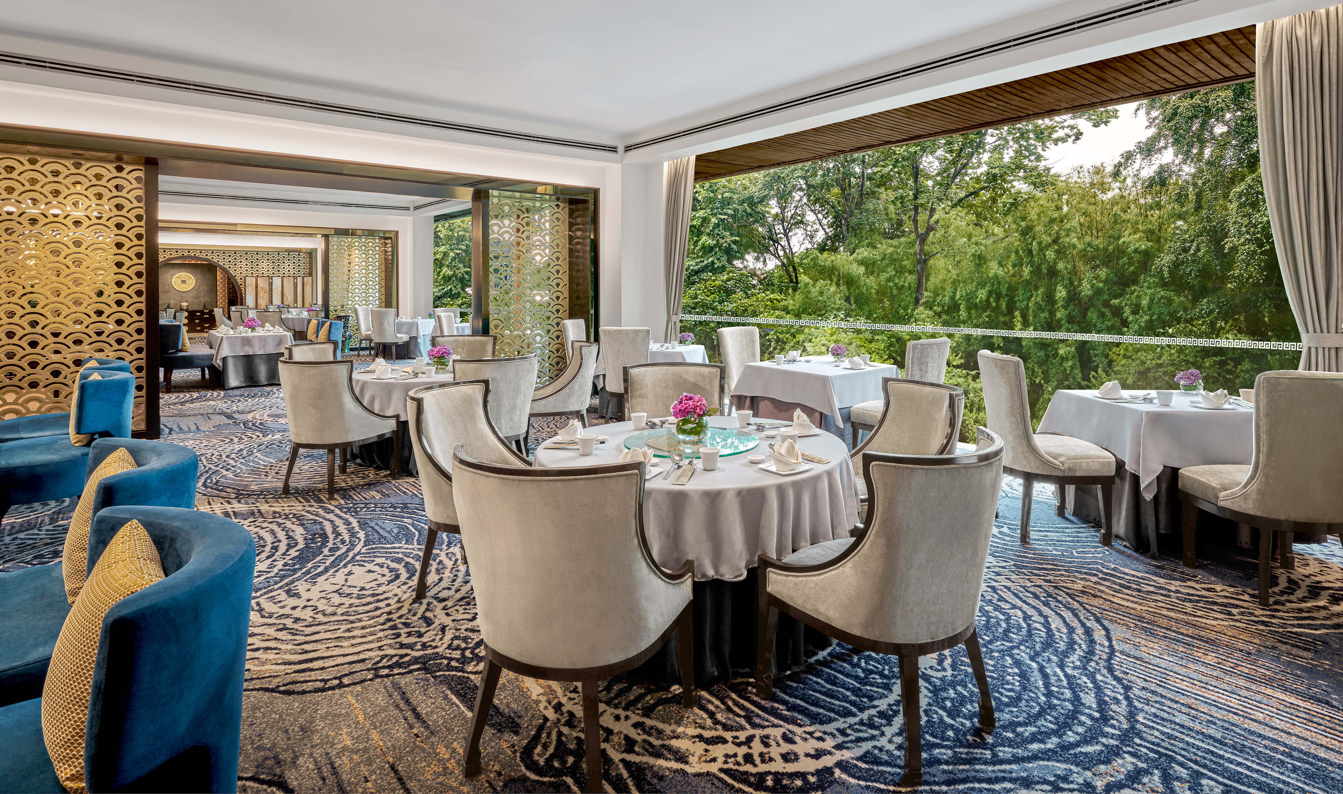 Elegant restaurant interior with round tables, gold screens, and lush tree view.