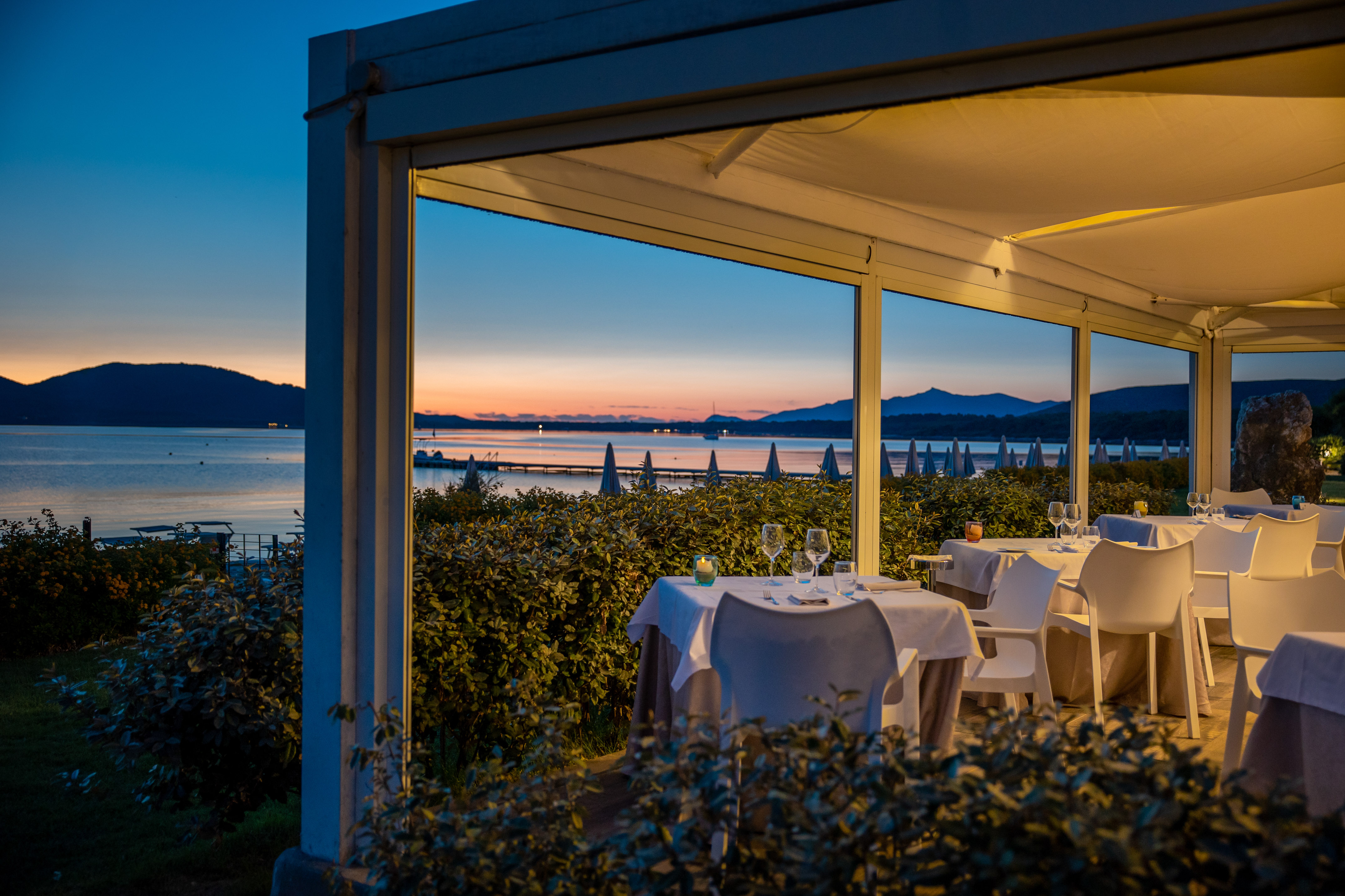 Outdoor restaurant tables with lit canopy, overlooking sea and dusk sky.