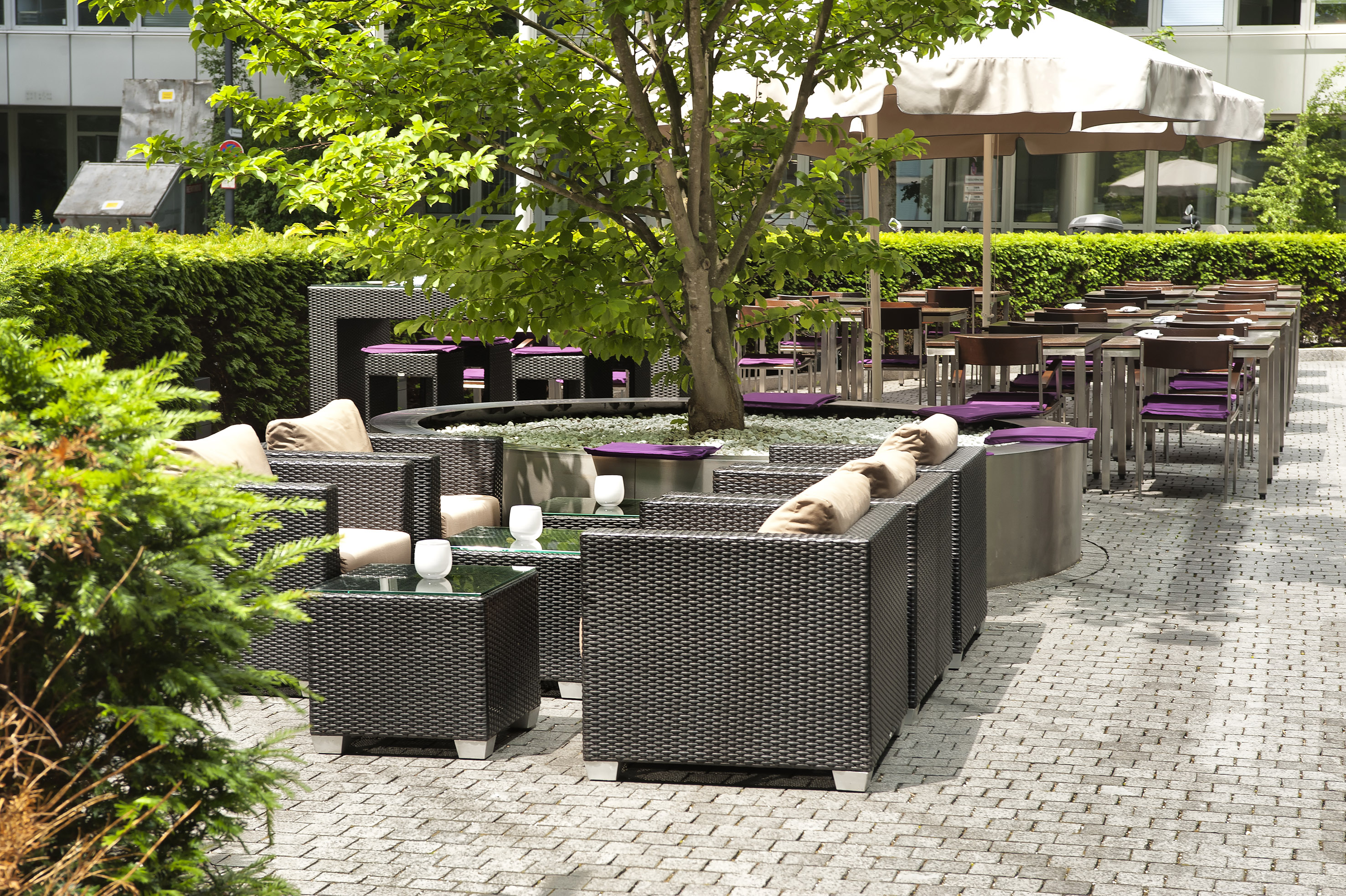 Modern outdoor patio with wicker lounge seating, cafe tables, and a central tree.