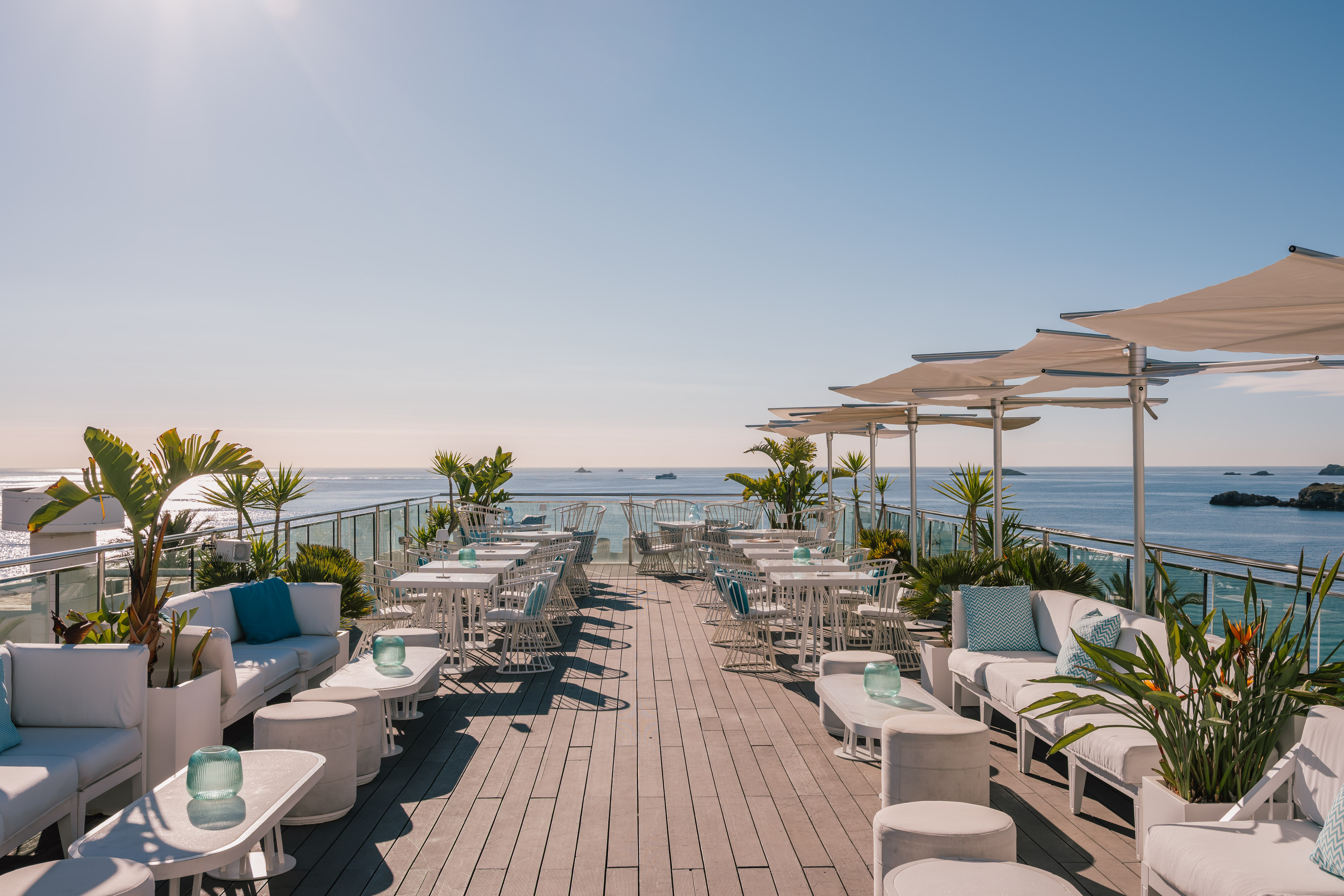 Rooftop lounge bar with white outdoor seating, sun umbrellas, and ocean views.