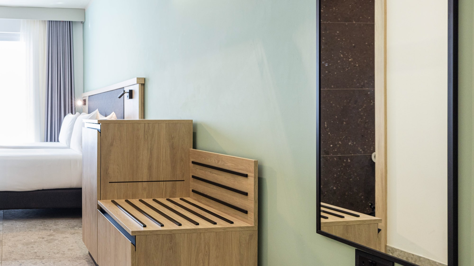 Modern hotel room with light green wall, wooden luggage rack, hangers, and mirror.