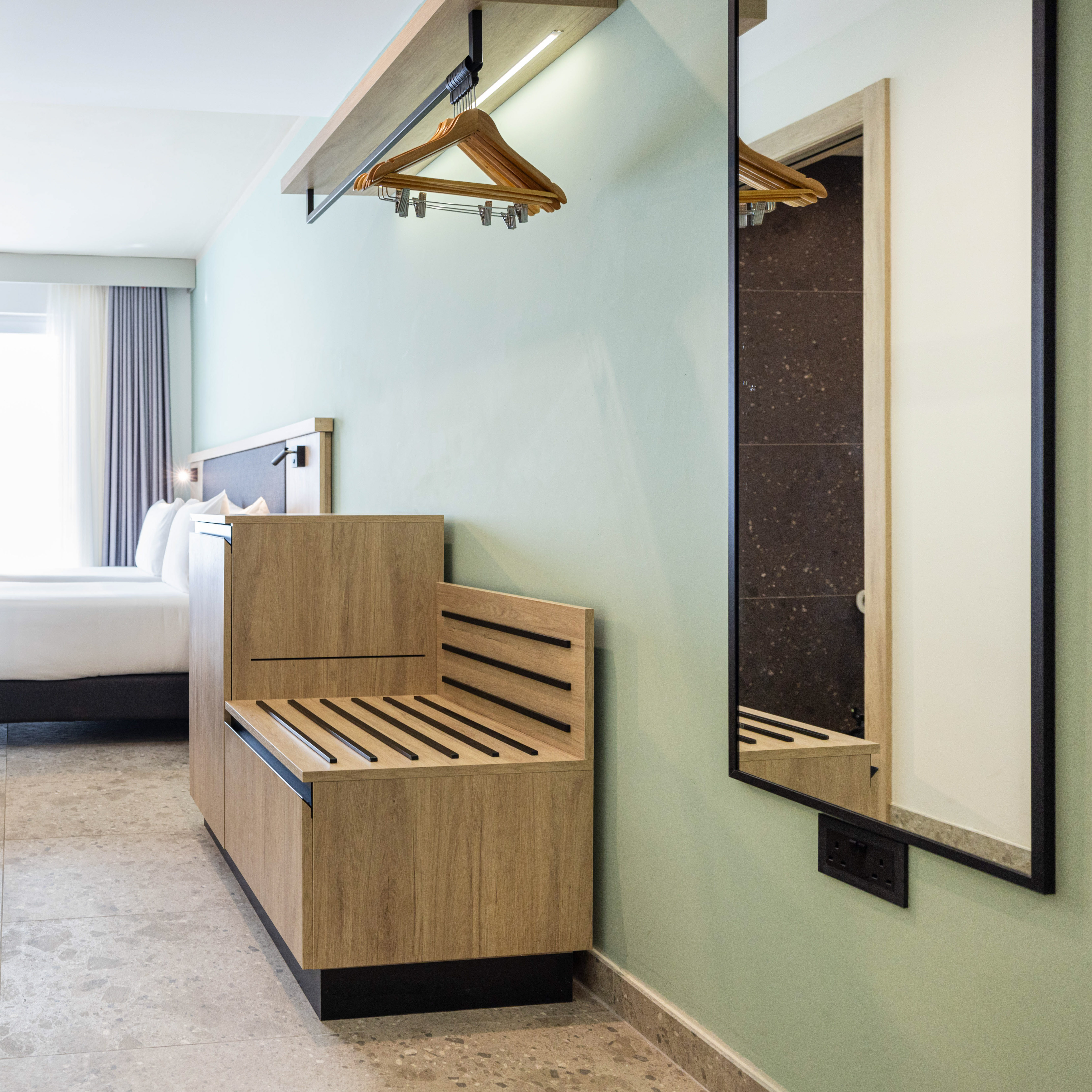 Modern hotel room with light green wall, wooden luggage rack, hangers, and mirror.