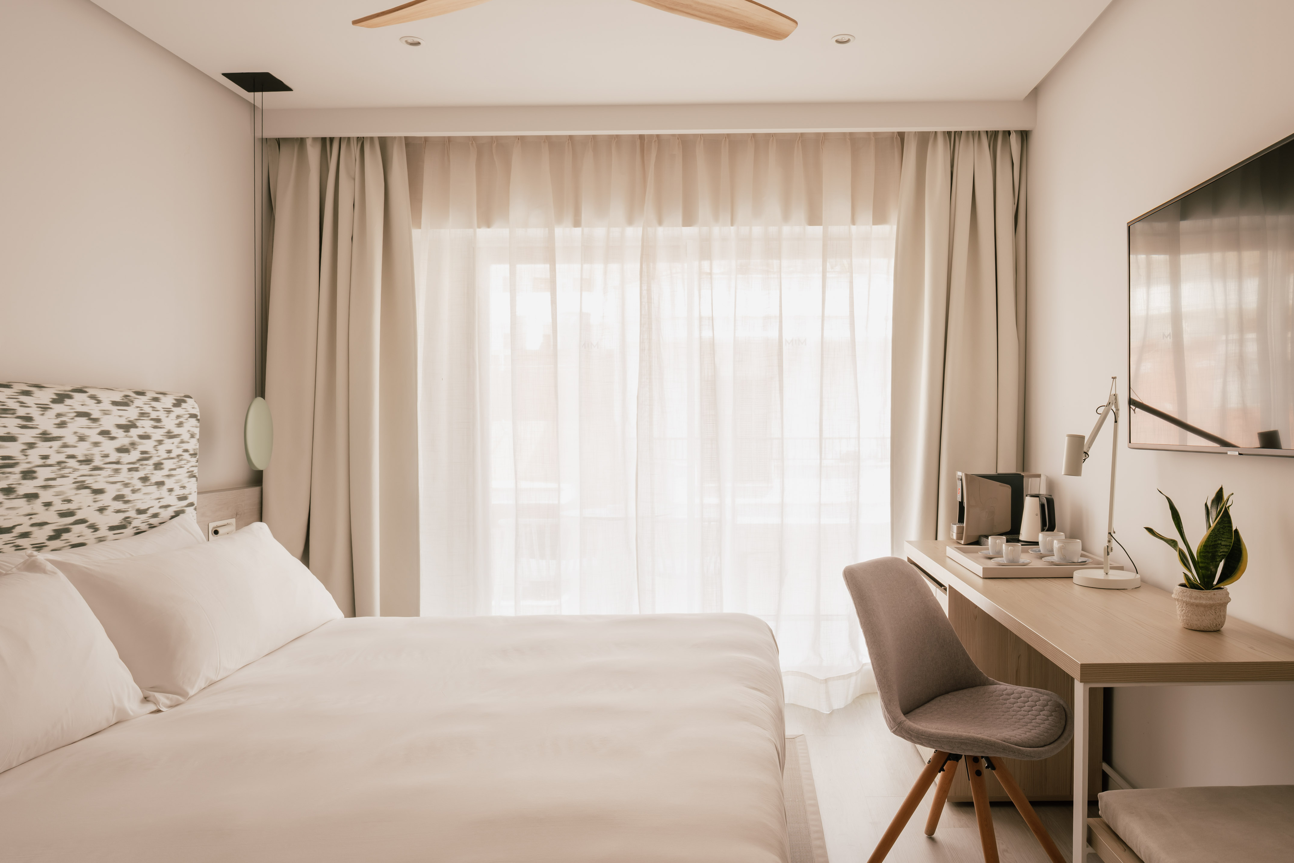 Dormitorio de hotel moderno con cama, escritorio, TV y ventana soleada.