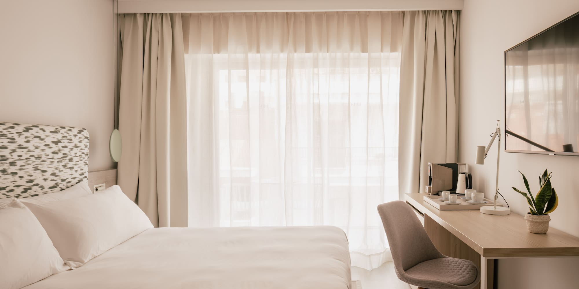 Modern hotel room with white bed, desk, coffee maker, and bright window.