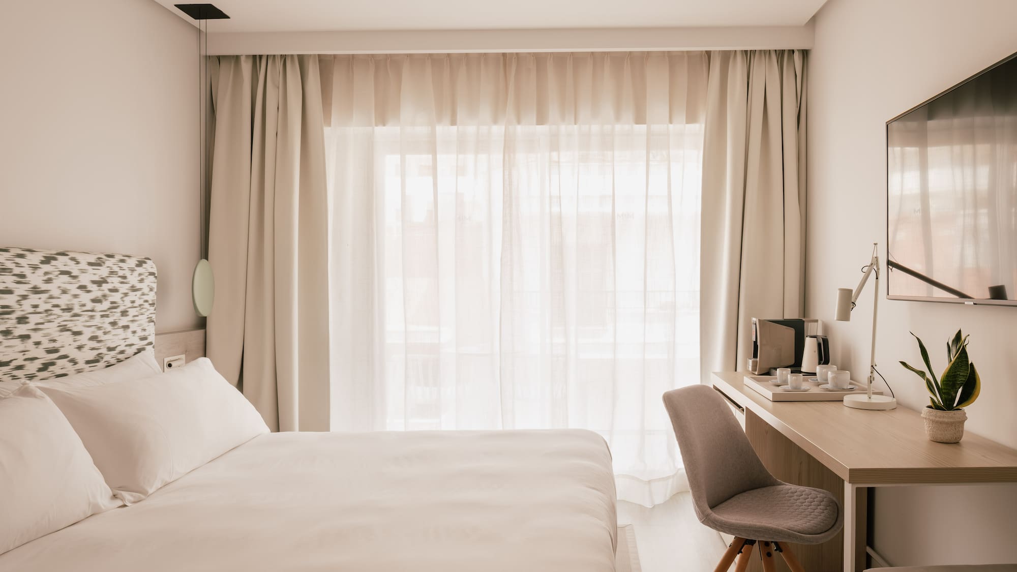 Modern hotel room with white bed, desk, coffee maker, and bright window.