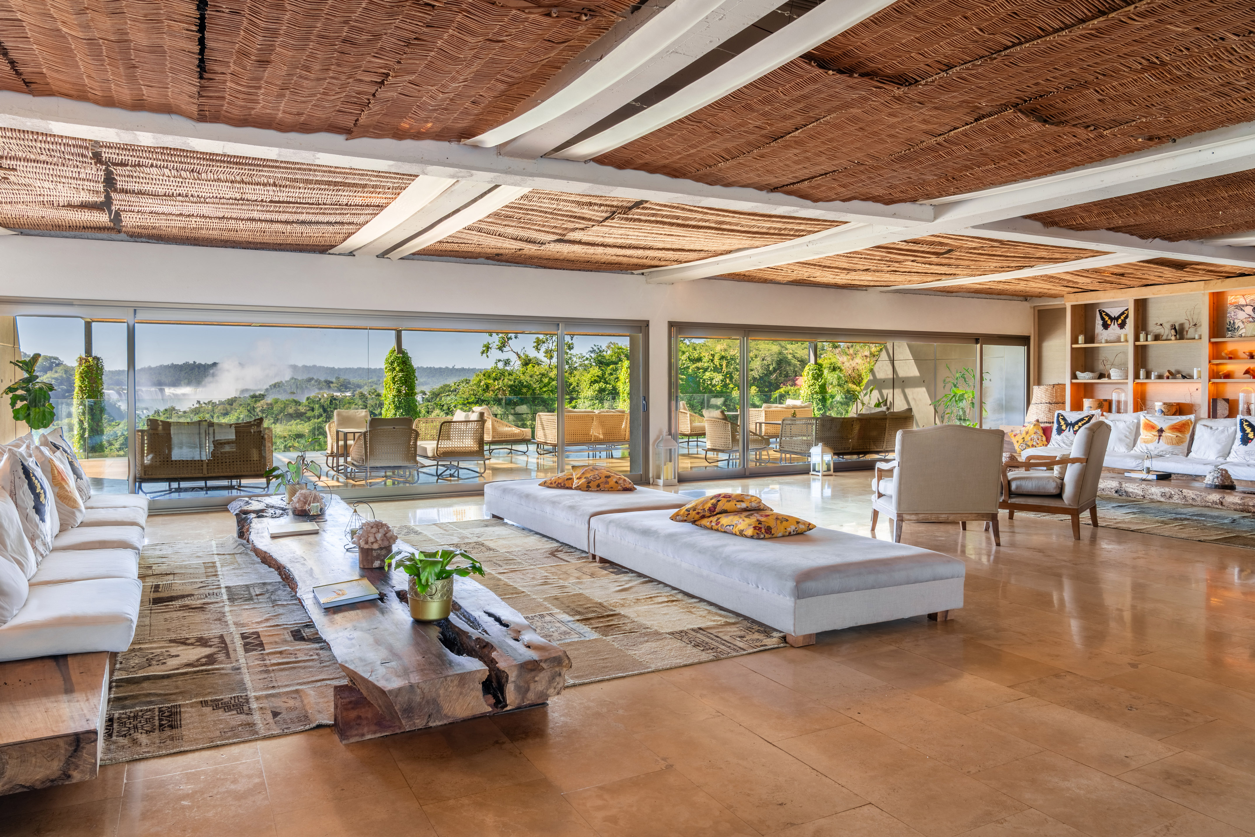 Lounge with panoramic waterfall view, woven ceiling, and rustic wood table.