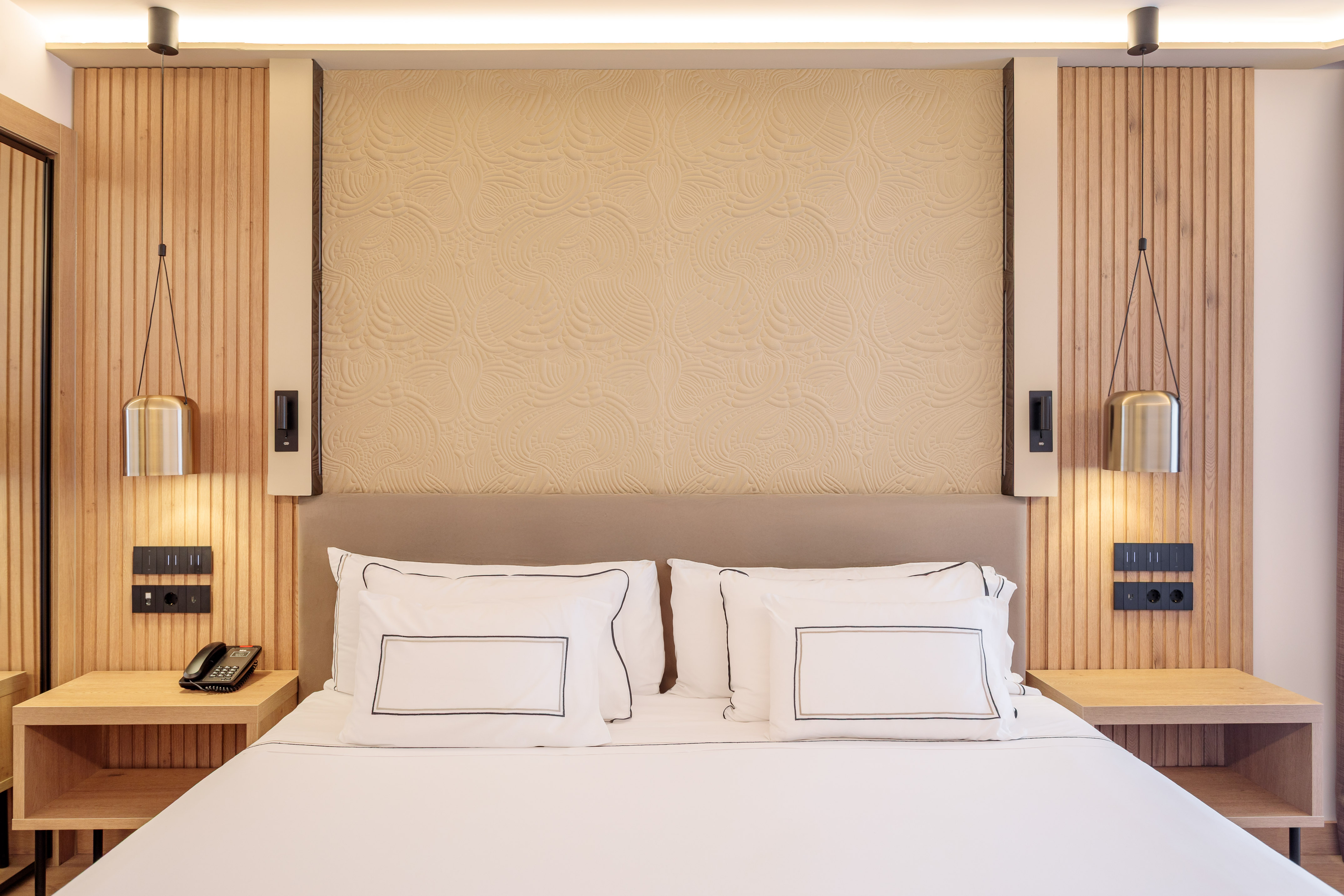 Modern hotel room with king bed, wood paneling, textured wall, and pendant lights.