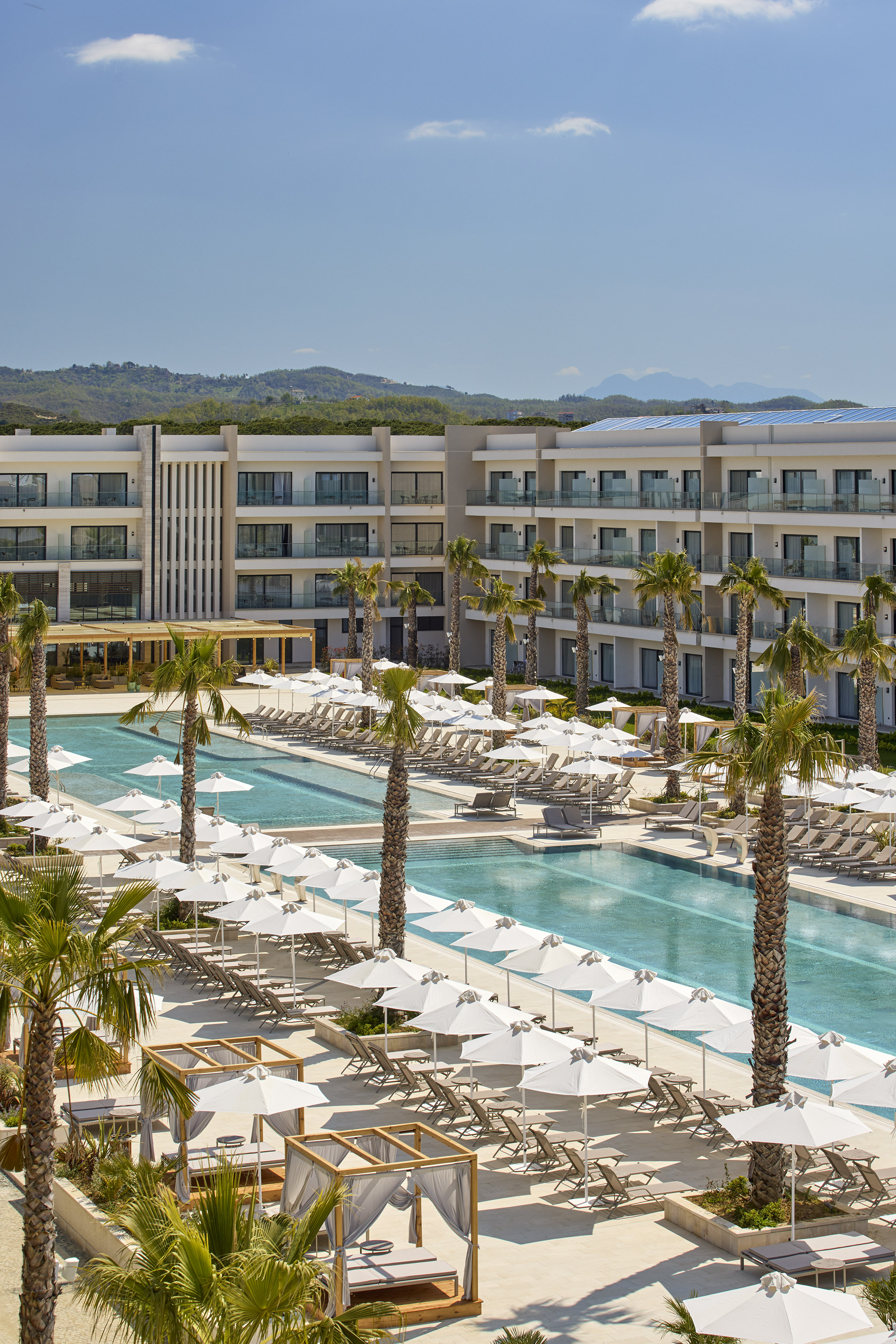 Modern resort hotel with large pools, palm trees, loungers, and white umbrellas.