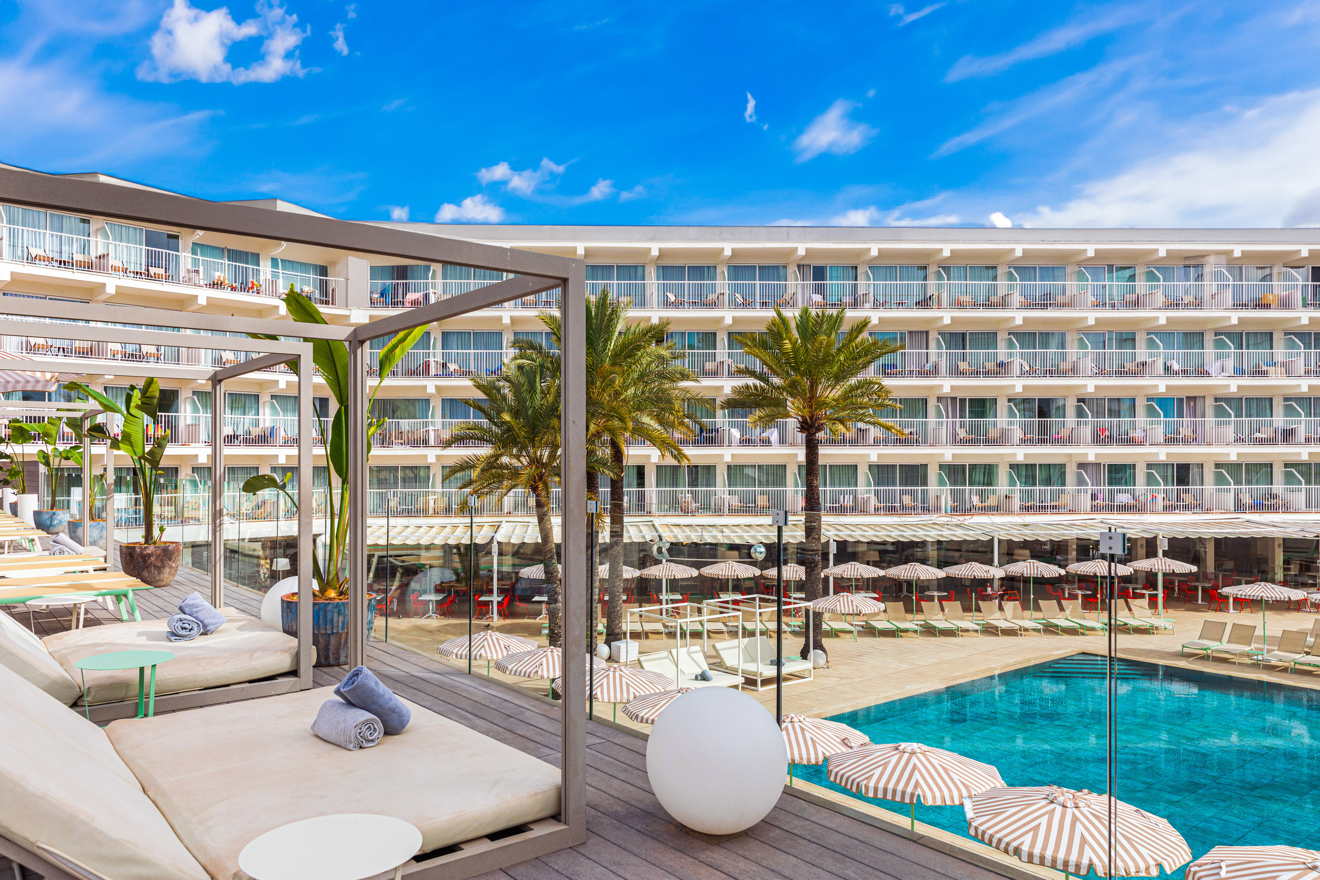 Sunny resort hotel with swimming pool, sunbeds, palm trees, and balconies.