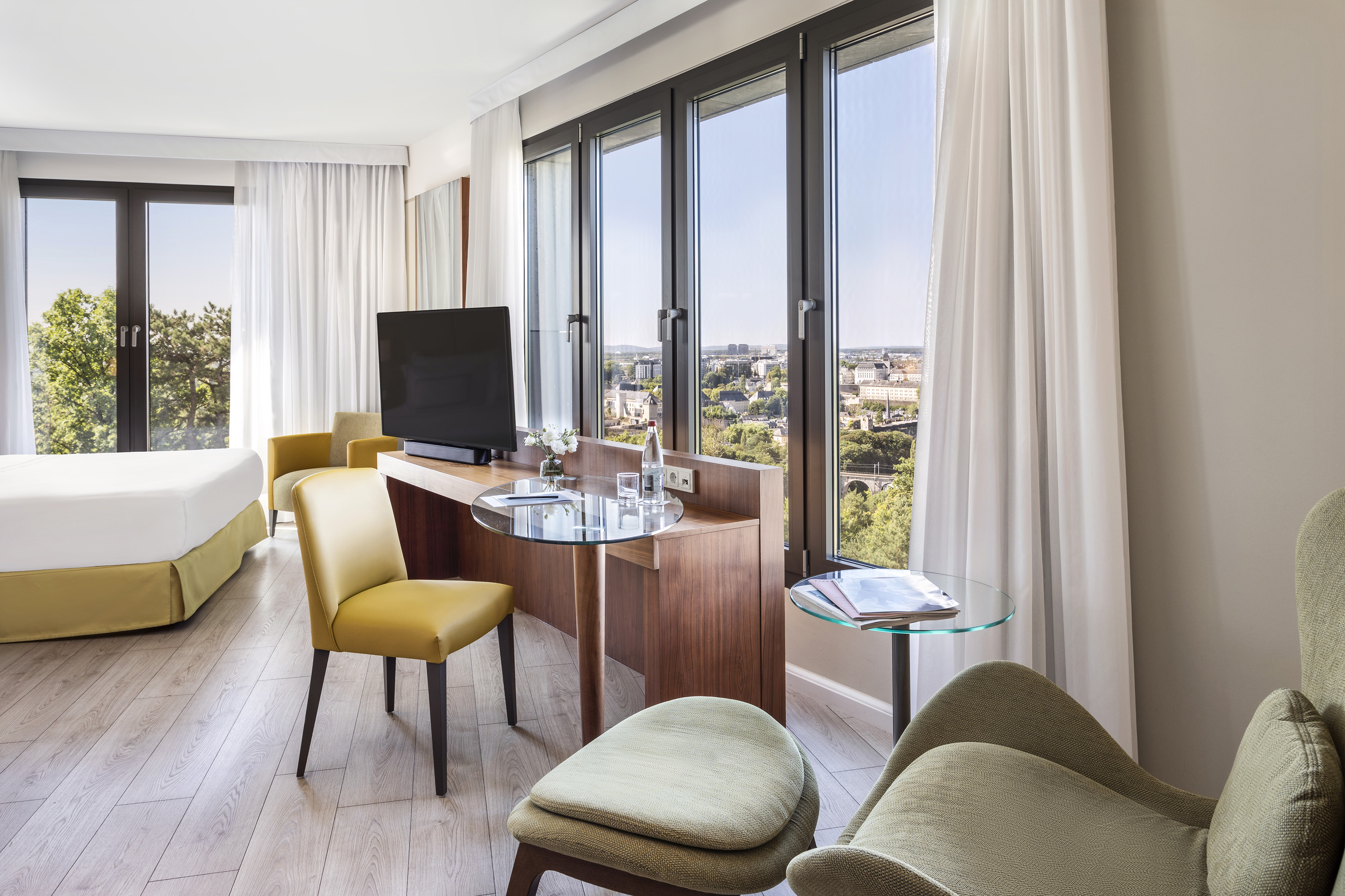 Modern hotel room with panoramic city view, desk, TV, and comfortable chairs.