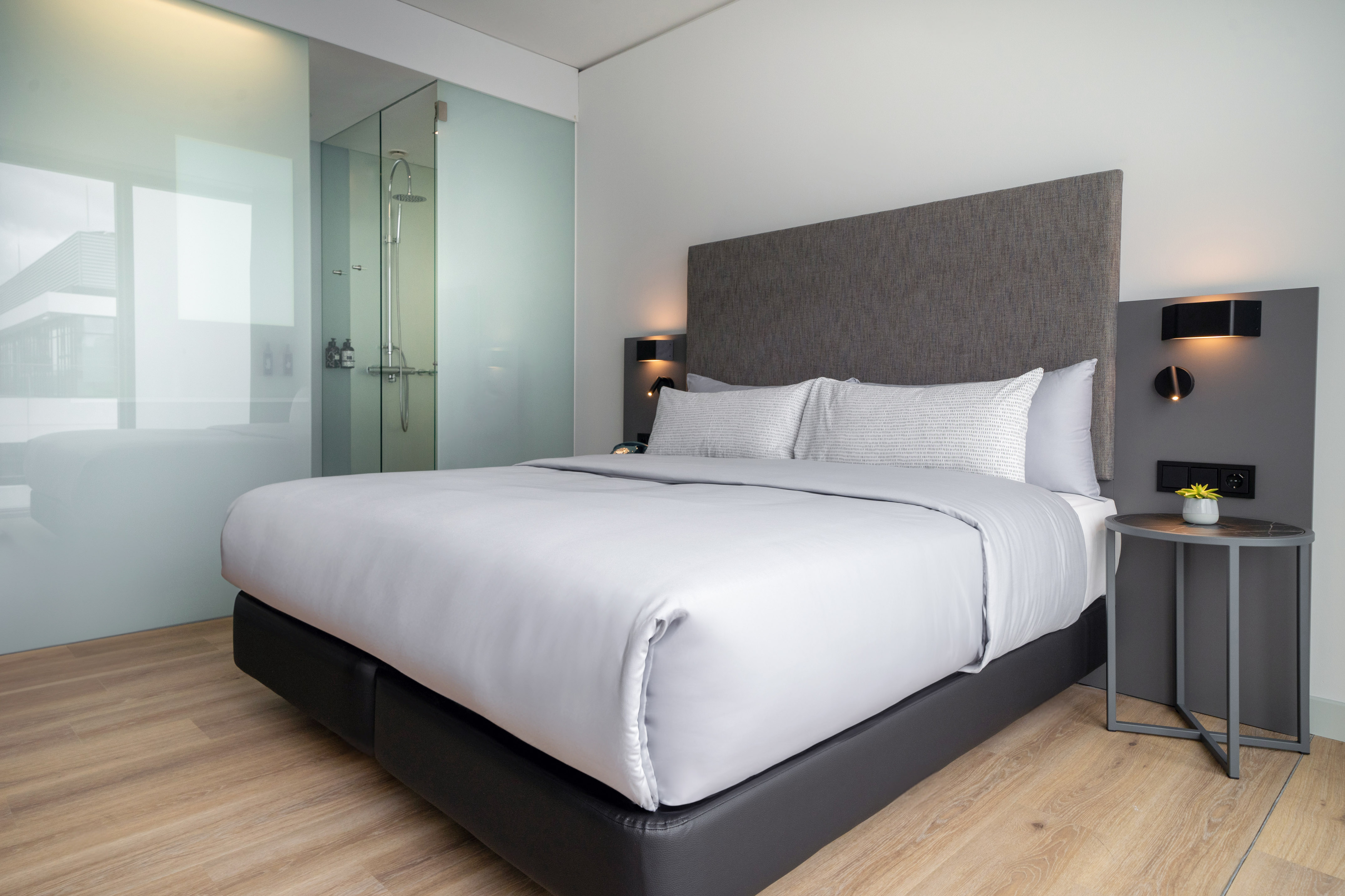 Modern hotel room with king bed, grey bedding, wood floor, and glass shower partition.