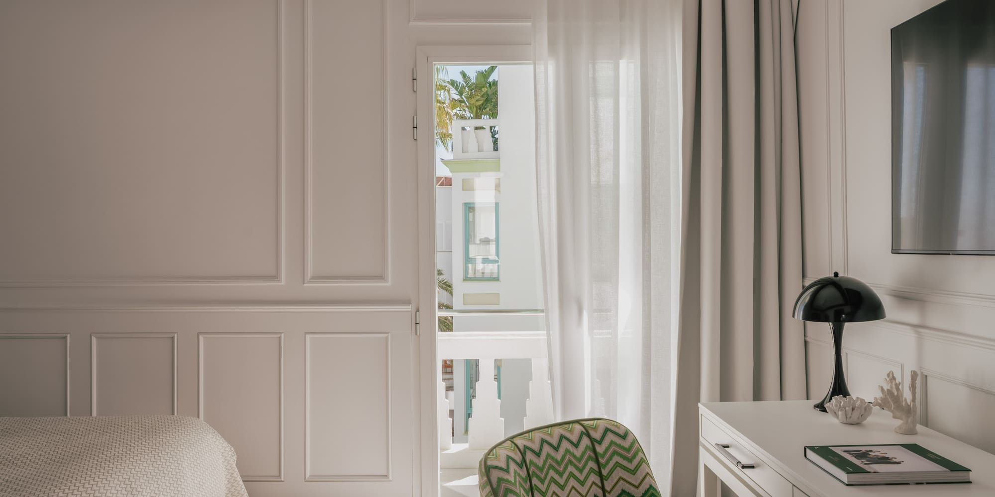 Modern hotel bedroom: white paneled walls, green chevron chair, balcony view.