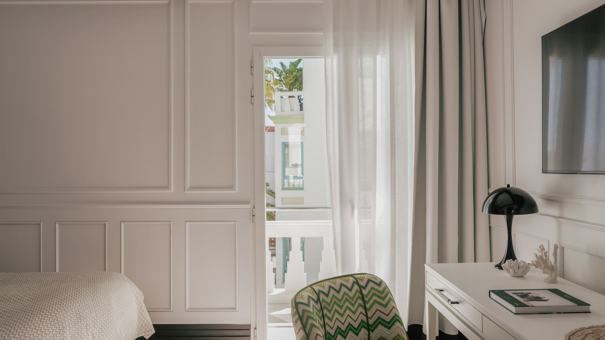 Modern white bedroom with balcony view, chevron chair, and desk.