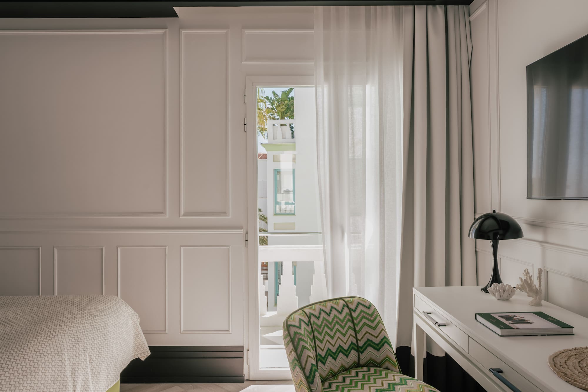 Modern hotel bedroom: white paneled walls, green chevron chair, balcony view.