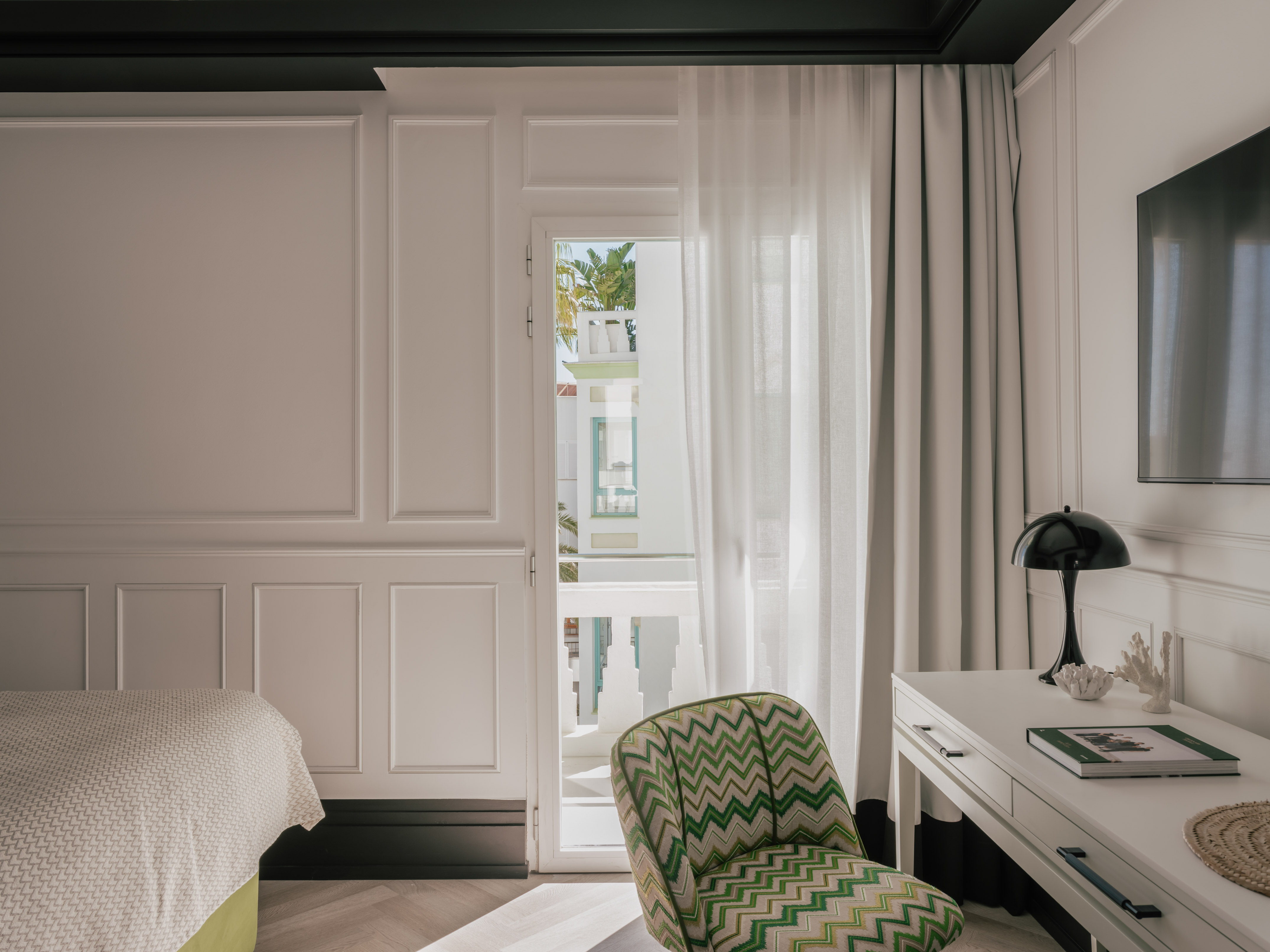 Modern hotel bedroom: white paneled walls, green chevron chair, balcony view.