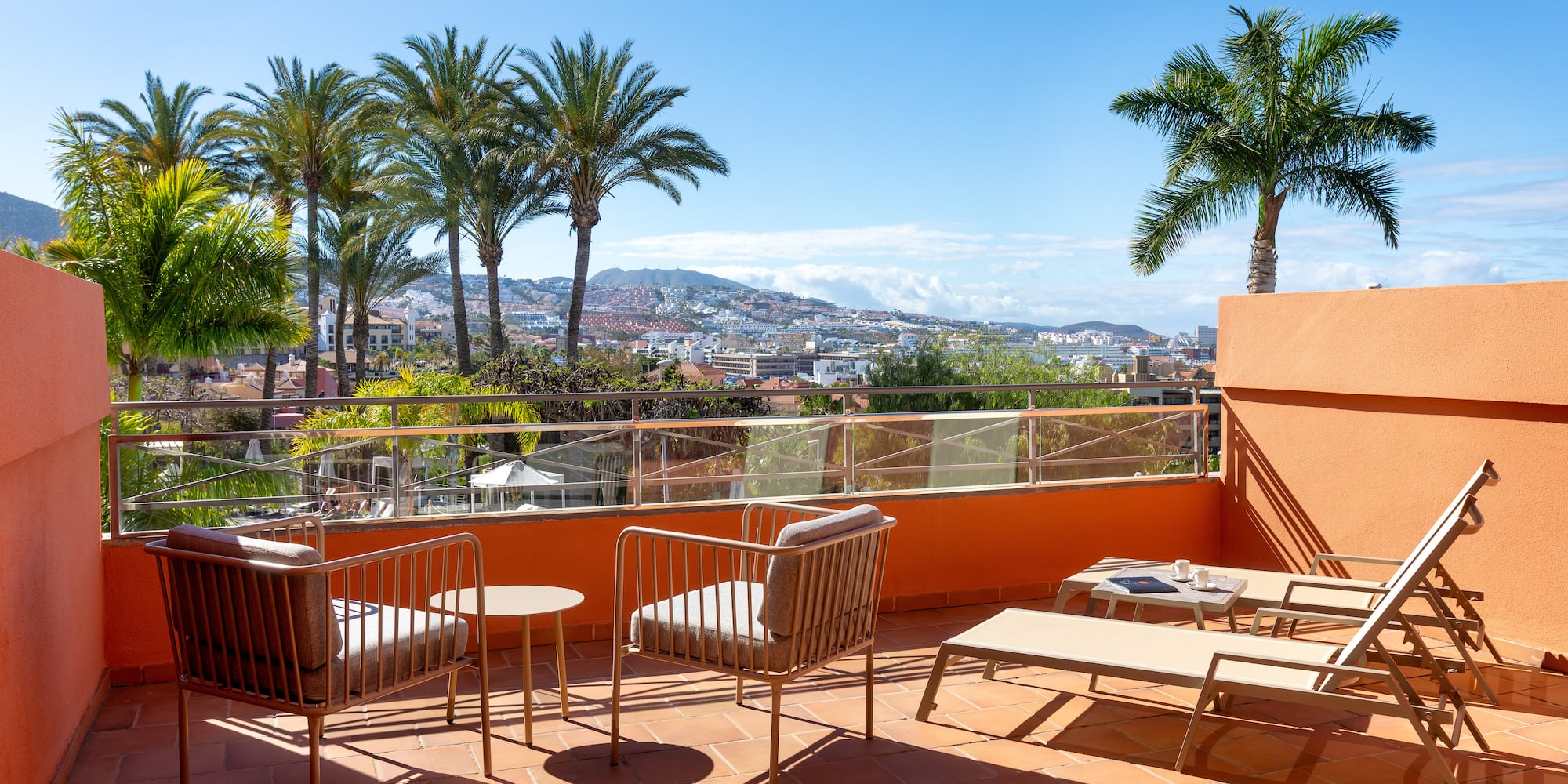 Sunny resort balcony with lounge chairs, palm trees, and city view.